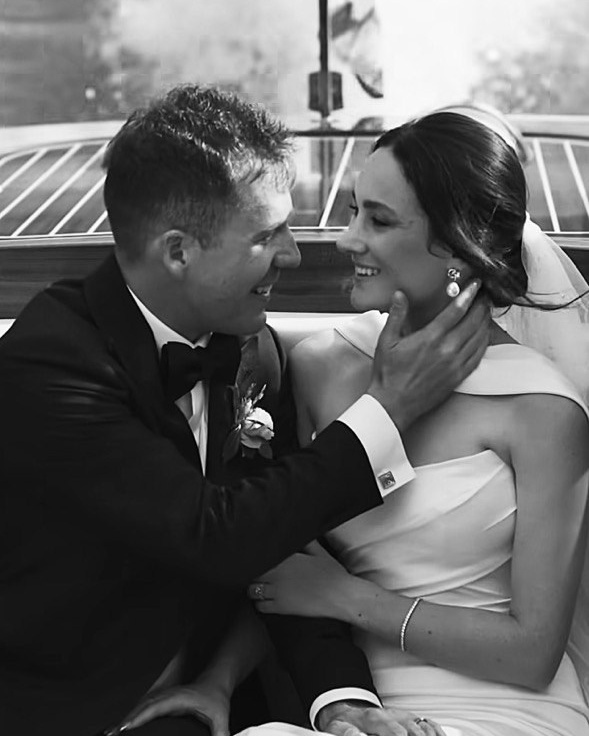A black-and-white photo of a bride standing on the beach with her back turned, holding her veil in the air as it flows in the wind, with the ocean in the background.
