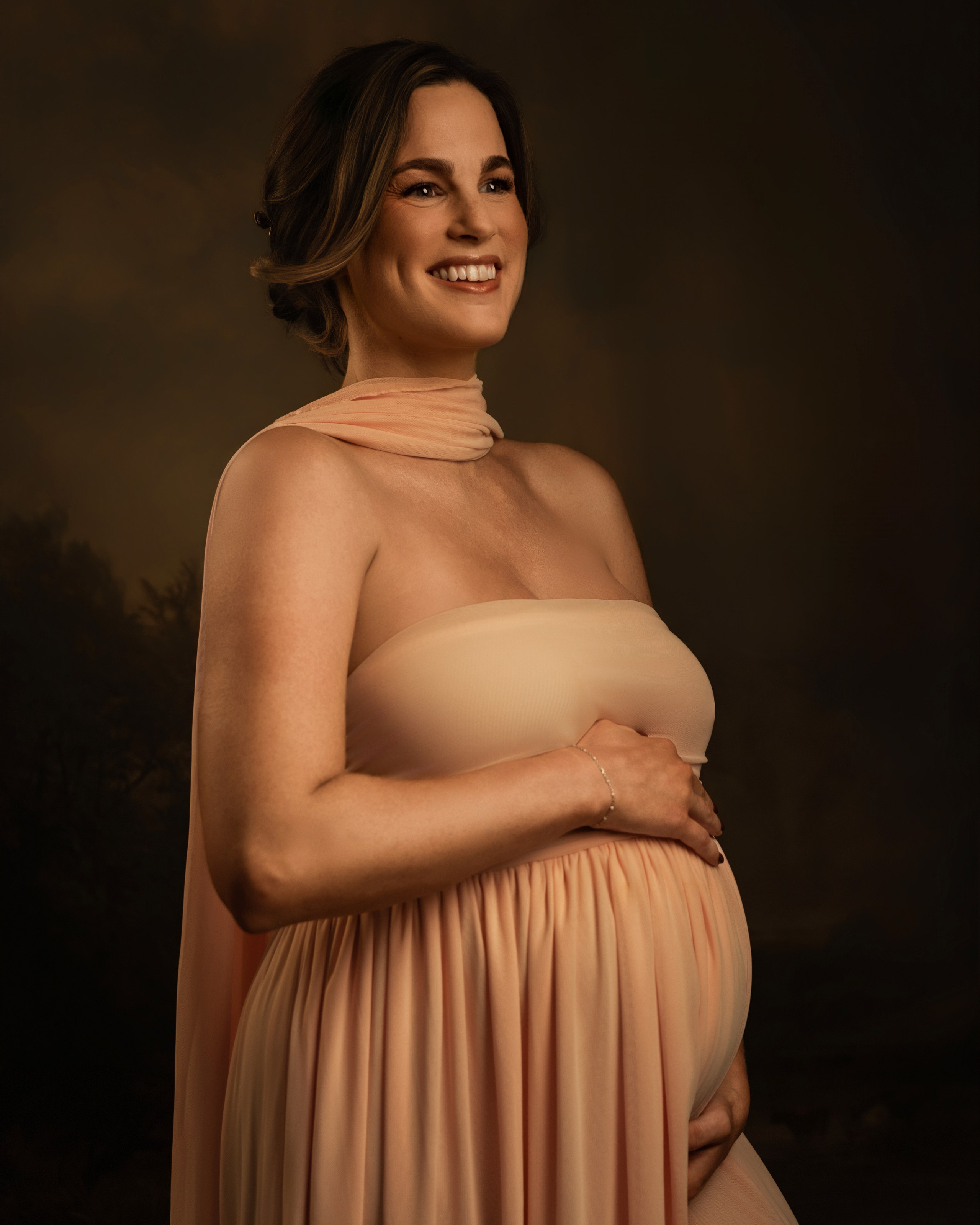 Pregnant woman in a soft blush gown, hands on her bump, dark backdrop