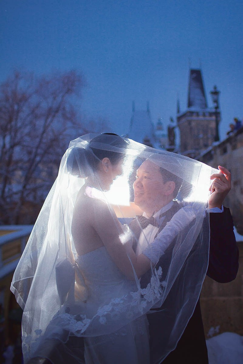 CY pulls Helen’s veil over his face near Charles Bridge at blue hour in Prague.