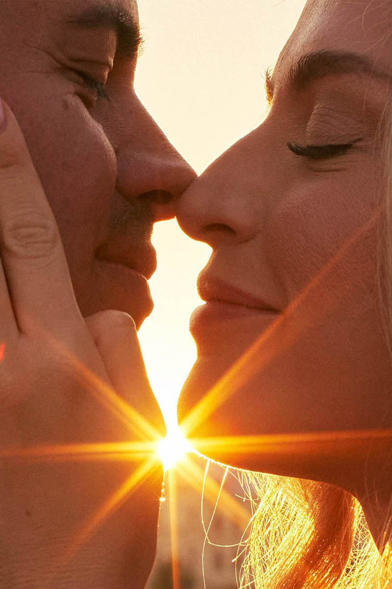 A gorgeous couple lovingly embraces as the sun flares vibrantly between their faces.