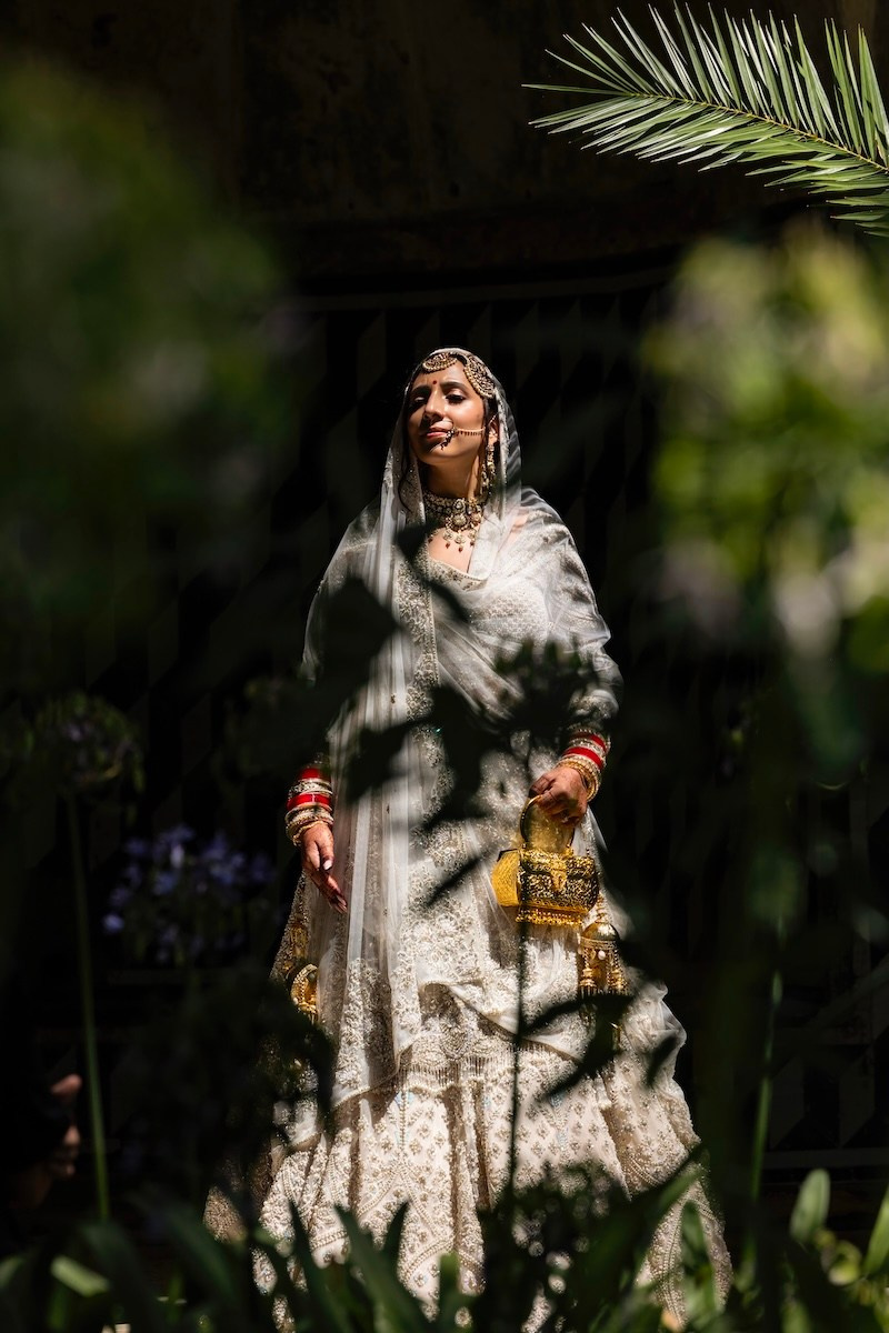 Indian bride in traditional bridal attire photographed through foliage in dramatic natural light in Portugal