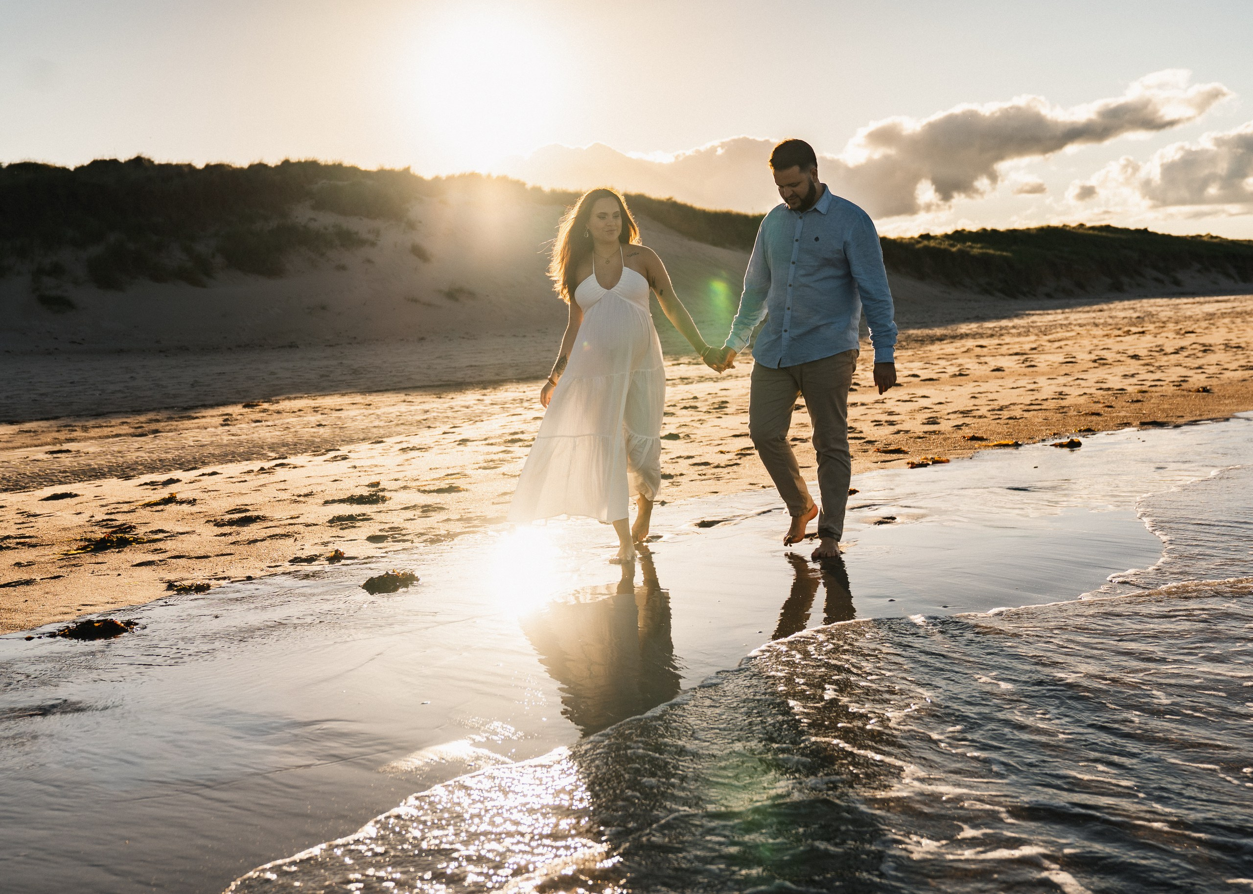 Pregnant couple walking hand in hand along the shoreline at sunset on Druridge Bay, Northumberland. Reflection in wet sand with dunes and golden light. Newcastle maternity photography.