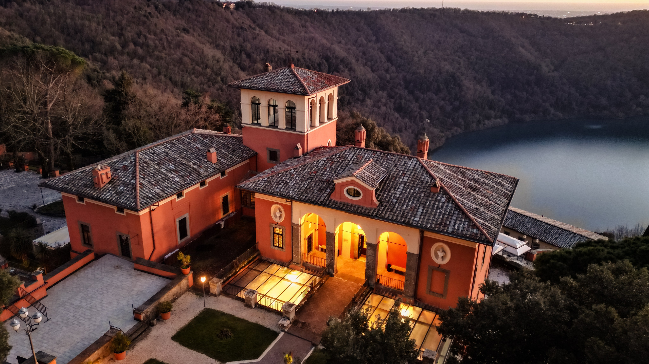 Vista aerea della splendida Villa del Cardinale illuminata la sera, con le sue facciate color terra e i tetti in tegole che si stagliano contro il paesaggio dei Castelli Romani e il lago sullo sfondo.