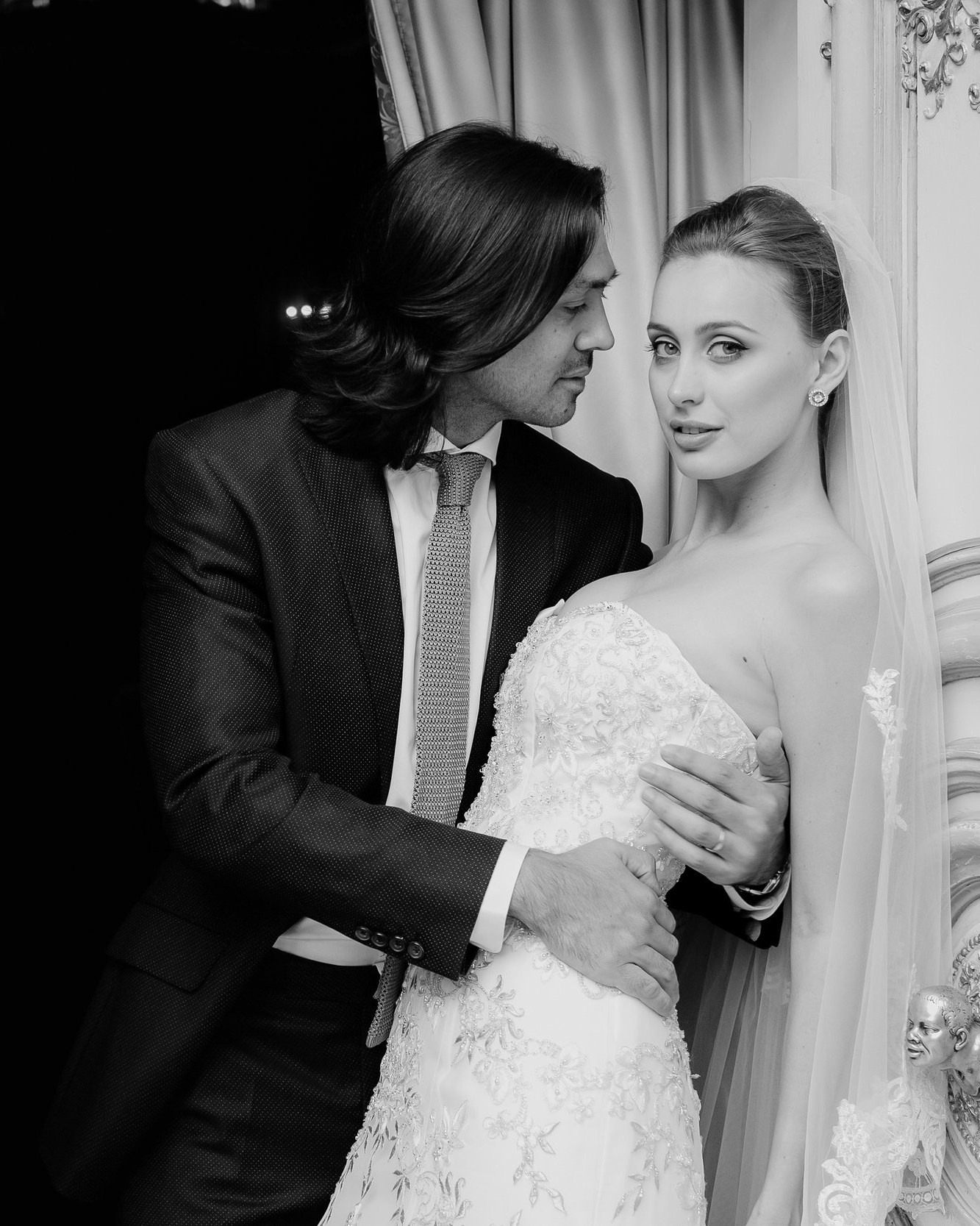 Fine art black-and-white wedding portrait: groom in a dark suit holds his bride in off-shoulder lace as they lean close near a sheer curtain