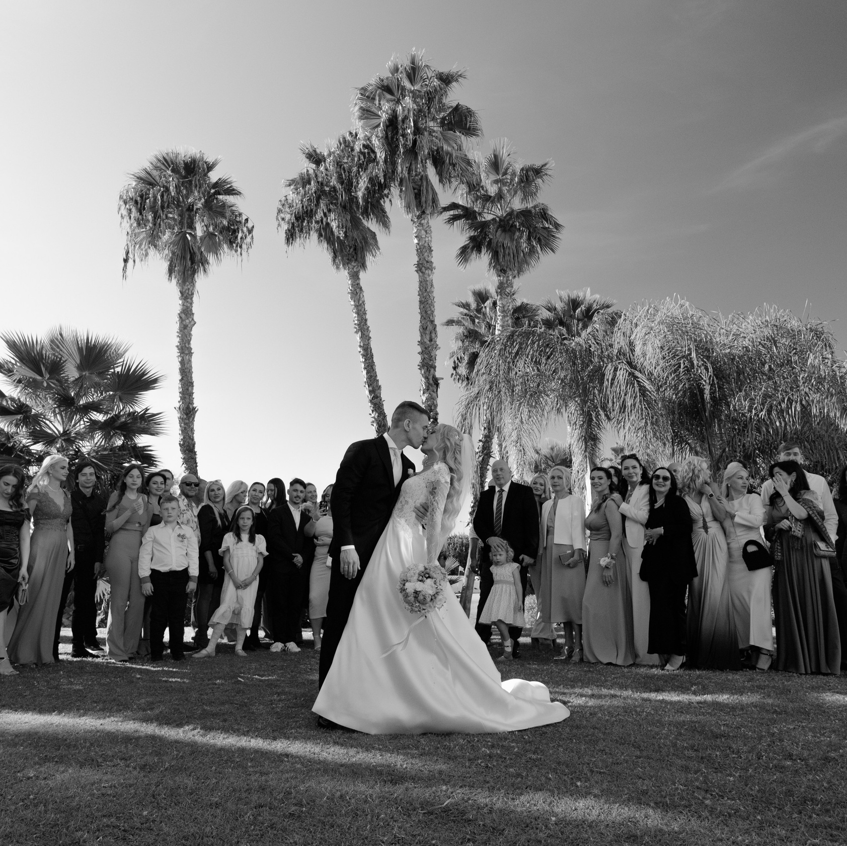 A group wedding photo with the bride and groom sharing a kiss in the foreground, surrounded by smiling guests, with tall palm trees under a bright sky.