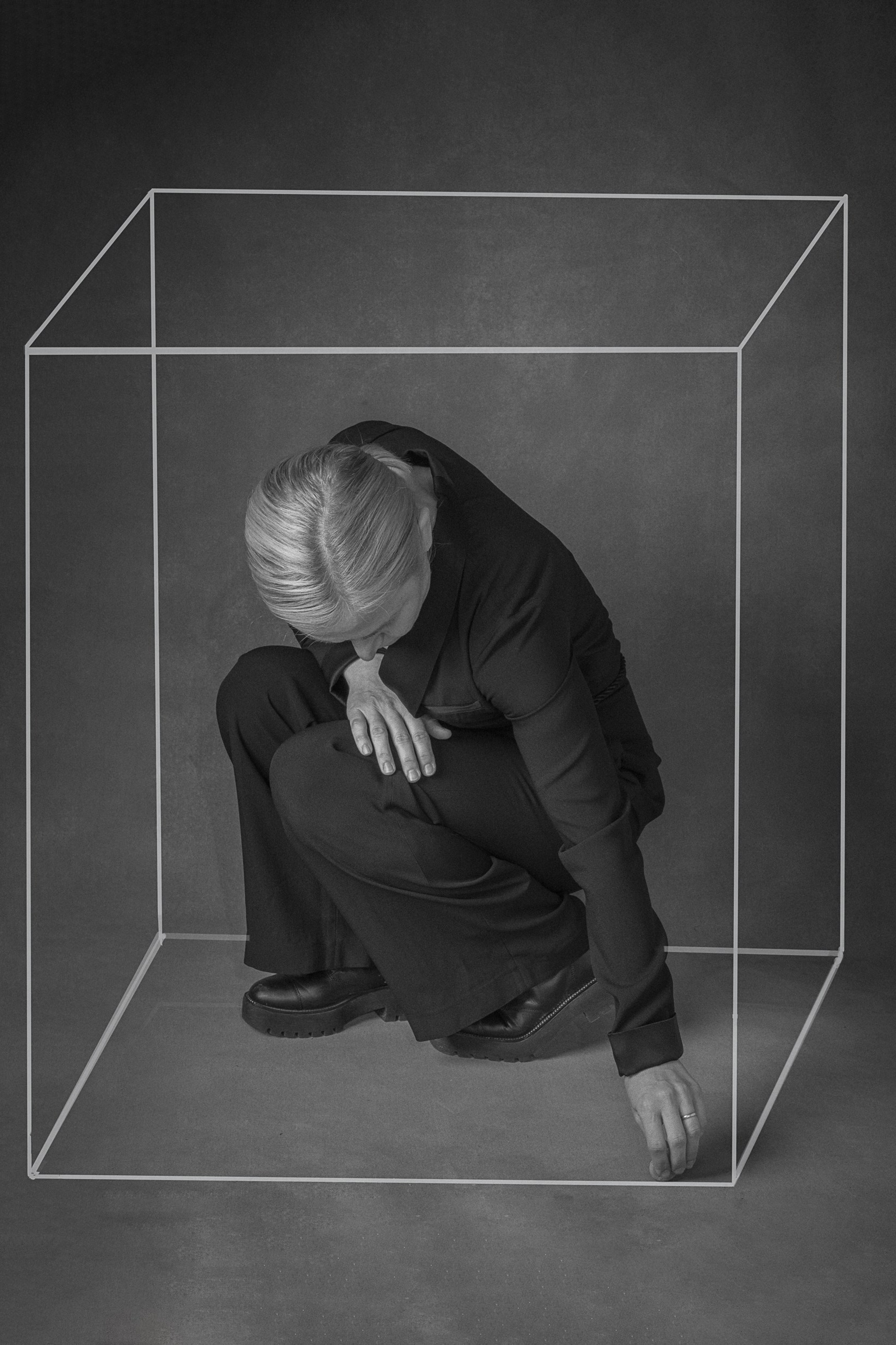 Black and white photograph of a person in a dark suit, crouched within a visually drawn cube.