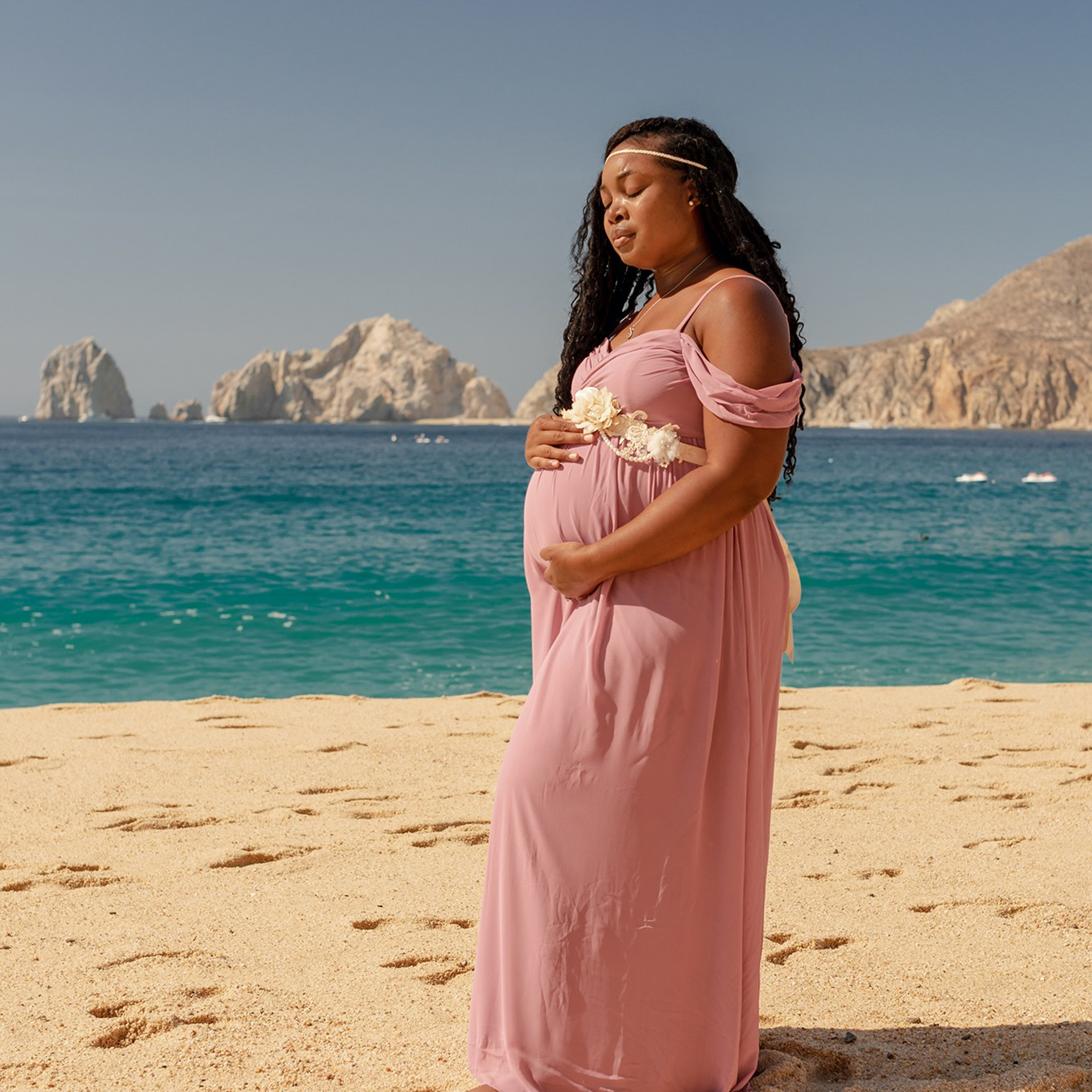 Maternity photoshoot in Los Cabos – pregnant woman in a flowing dress posing by the beach, portrait style with ocean waves and palm trees in the background