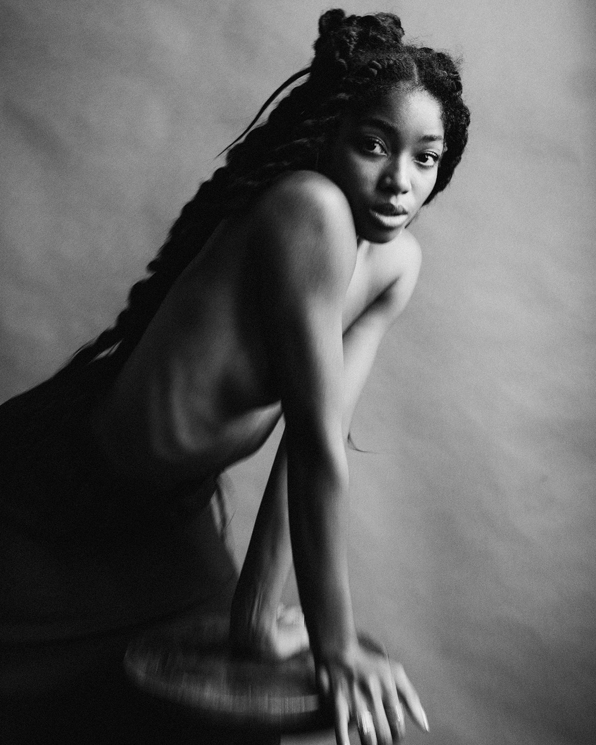 Black and white portrait of a woman with long braids leaning forward on a stool, gazing into the camera.