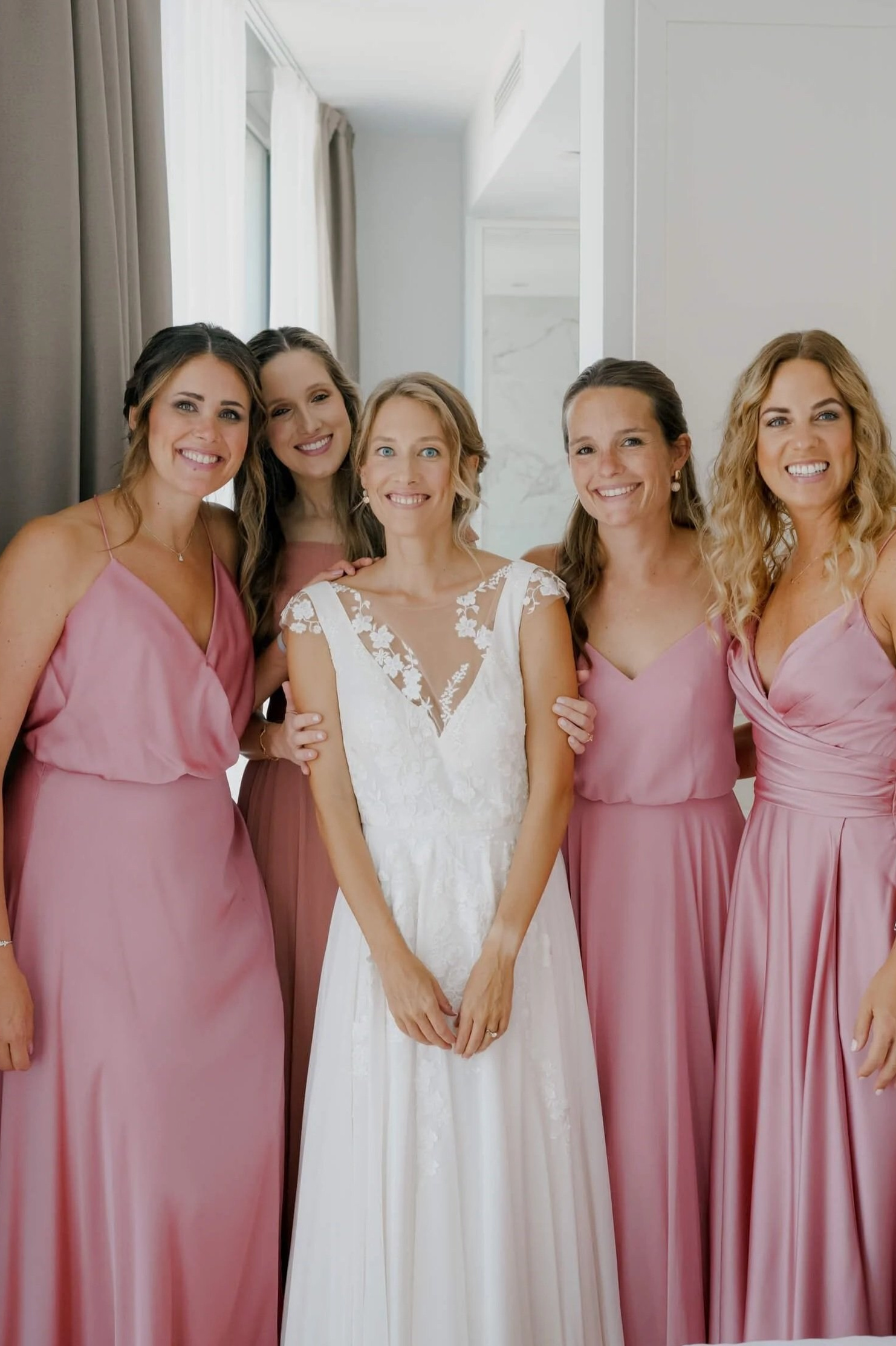 Bride with four bridesmaids in dusty pink gowns, bright window behind