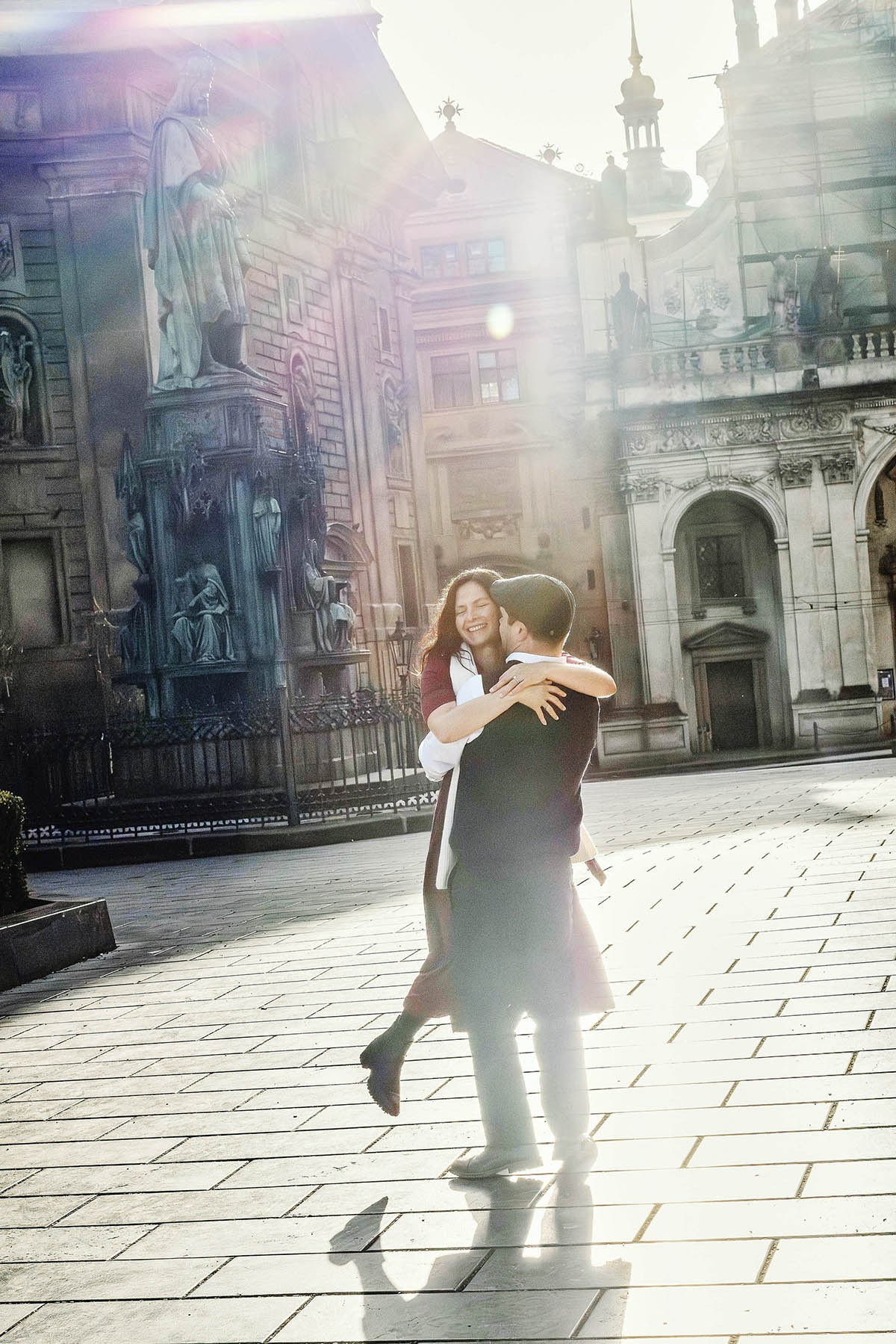 Woman smiling as she is lifted into the air beneath the statue of King Charles IV in Prague while golden sunrise light creates natural lens flare.