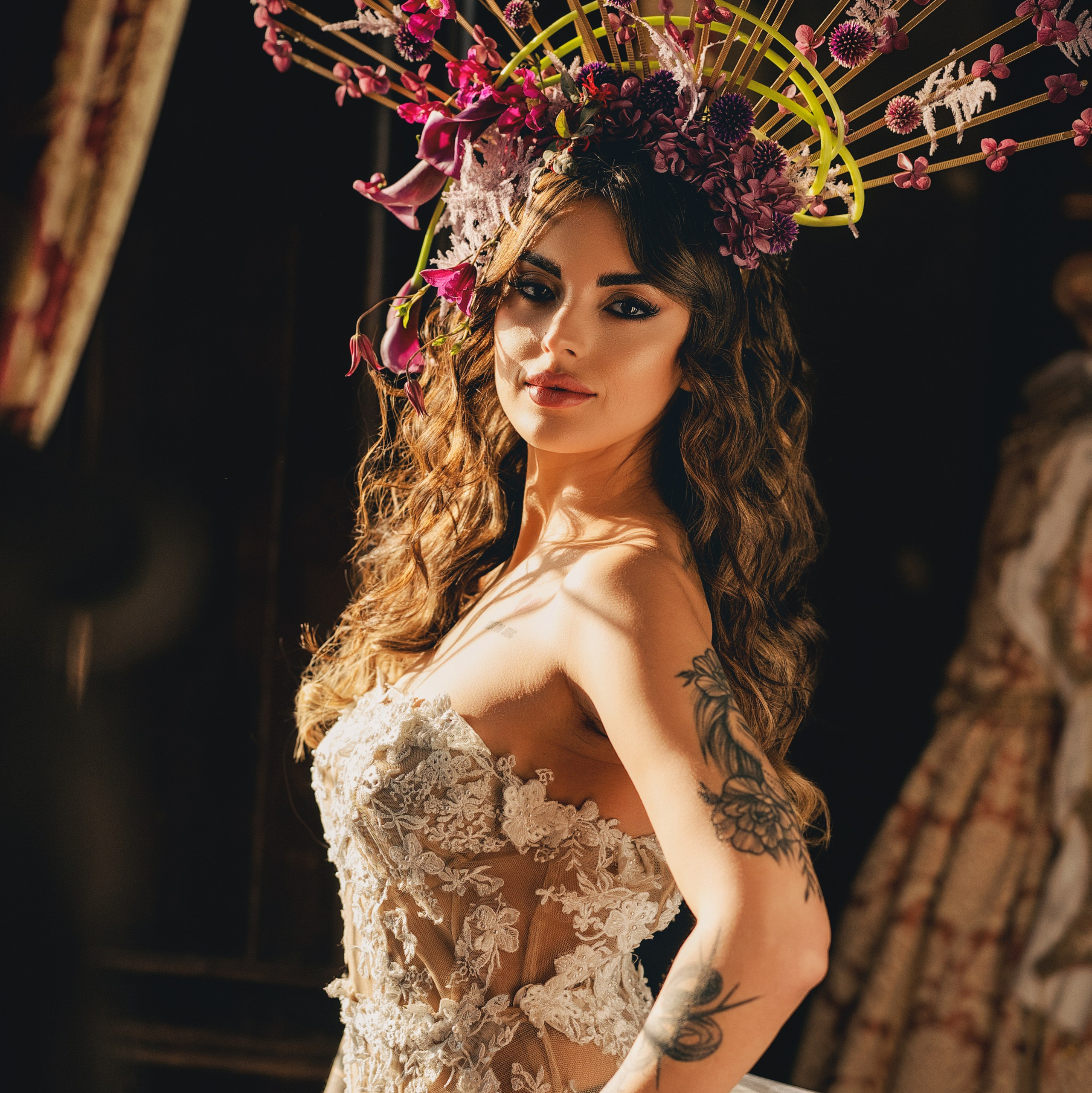 Sposa con acconciatura floreale in toni rosso e rosa indossa un abito da sposa in pizzo con schiena scoperta durante uno shooting di matrimonio artistico davanti ad una finestra