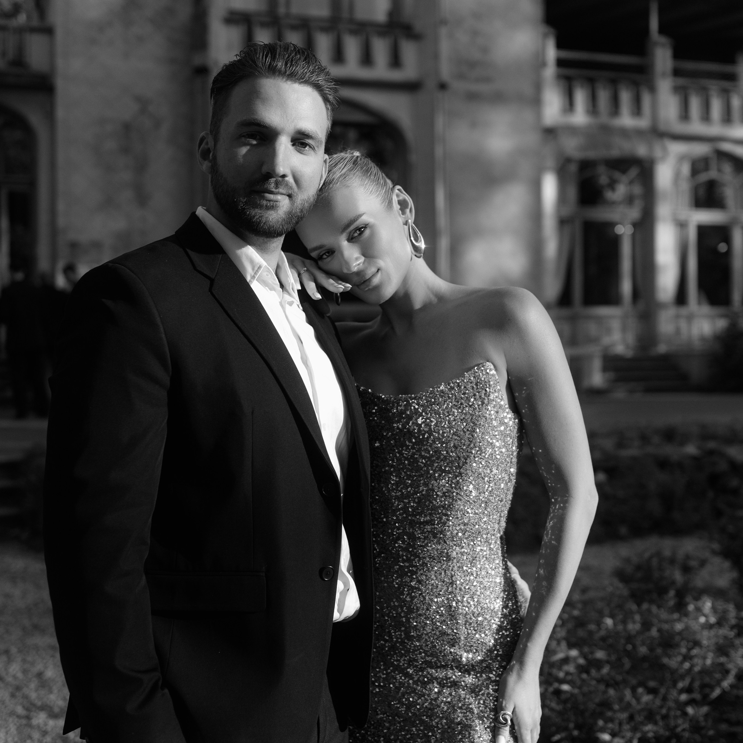 Black and white photo of a couple on the beach. A man in a light shirt gently hugs a woman in a long slit dress, they look calm and happy against the background of the sea and the horizon