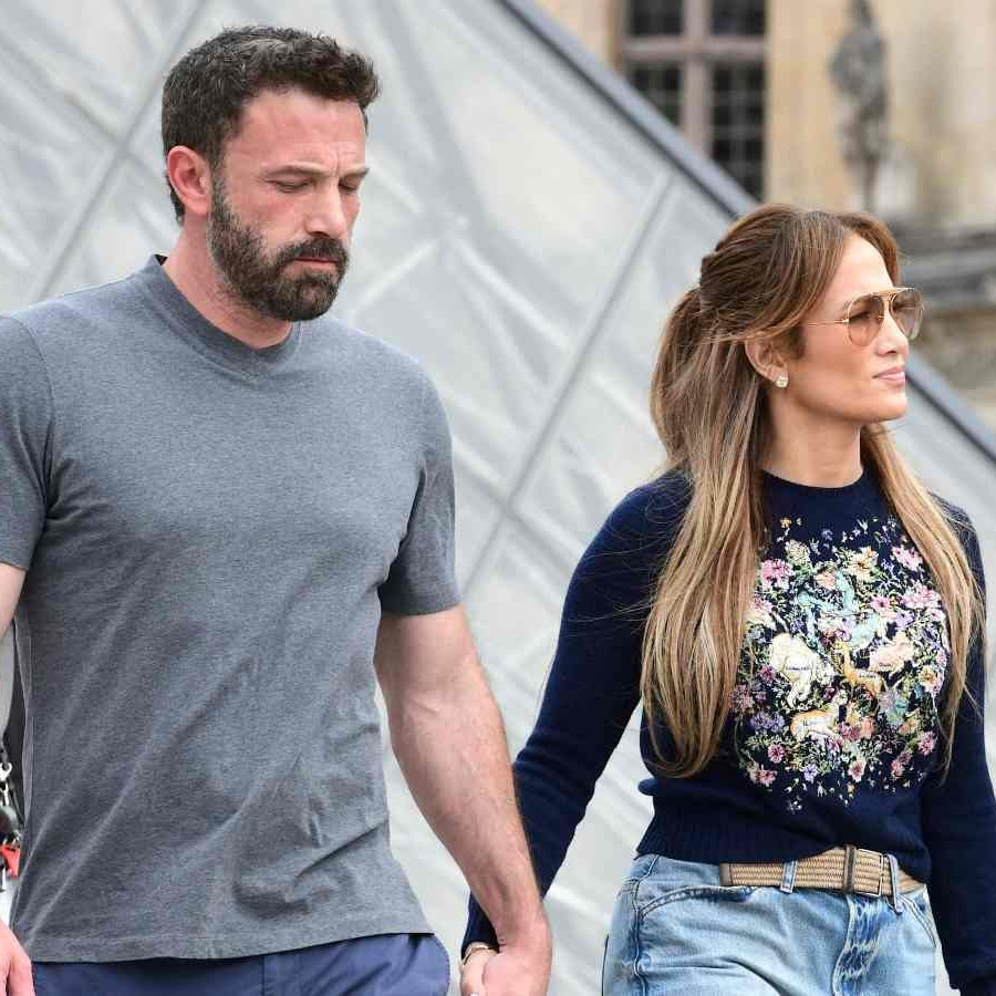 Бен Аффлек в серой футболке и Дженнифер Лопес в синем свитере с цветочным принтом и джинсах держатся за руки на фоне пирамиды Лувра, Париж 2024