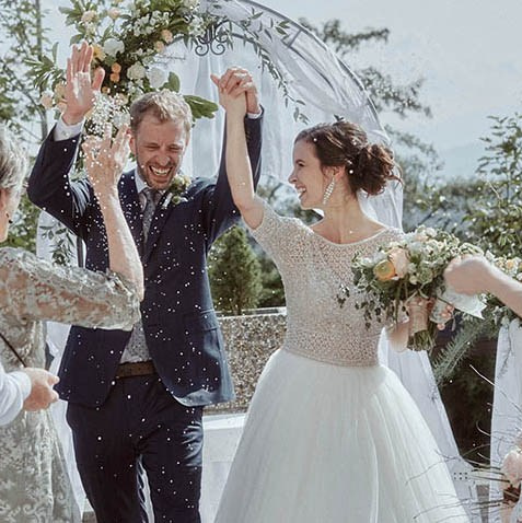 Red-haired bride holding her flowing veil aloft smiles seductively to her bouquet holding groom as he leans back against a railing in appreciation at the Vrtba Garden in Prague. 