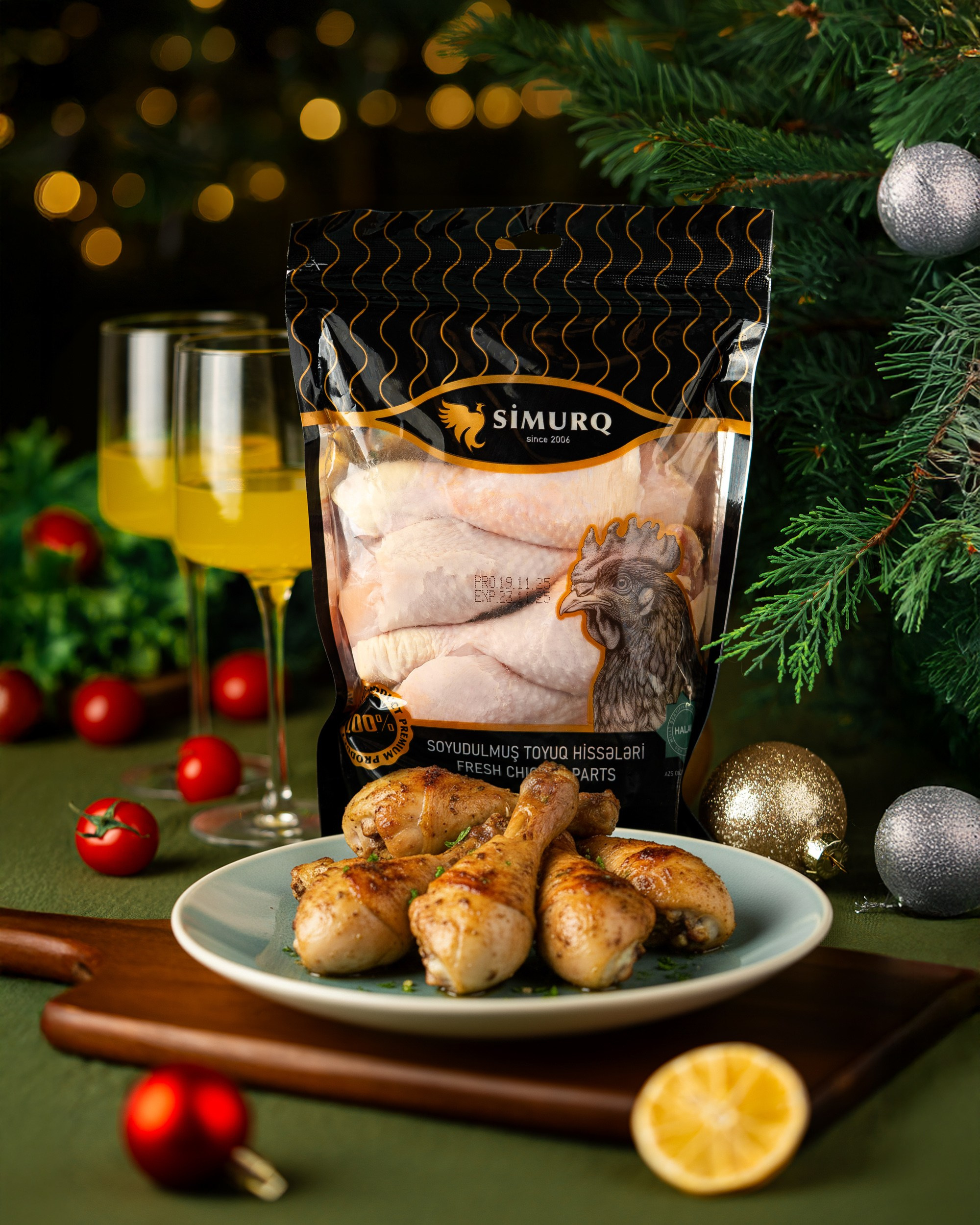 Yaşıl səth üzərində, Milad ağacı və parlayan fon işıqları ilə bəzədilmiş bir mühitdə. Mavi boşqabda qızardılmış dörd toyuq budu, arxasında isə "SİMURQ" markalı soyudulmuş toyuq budları (hissələri) paketi dayanır. Çerri pomidorları və limon dilimi ön planda yer alır.