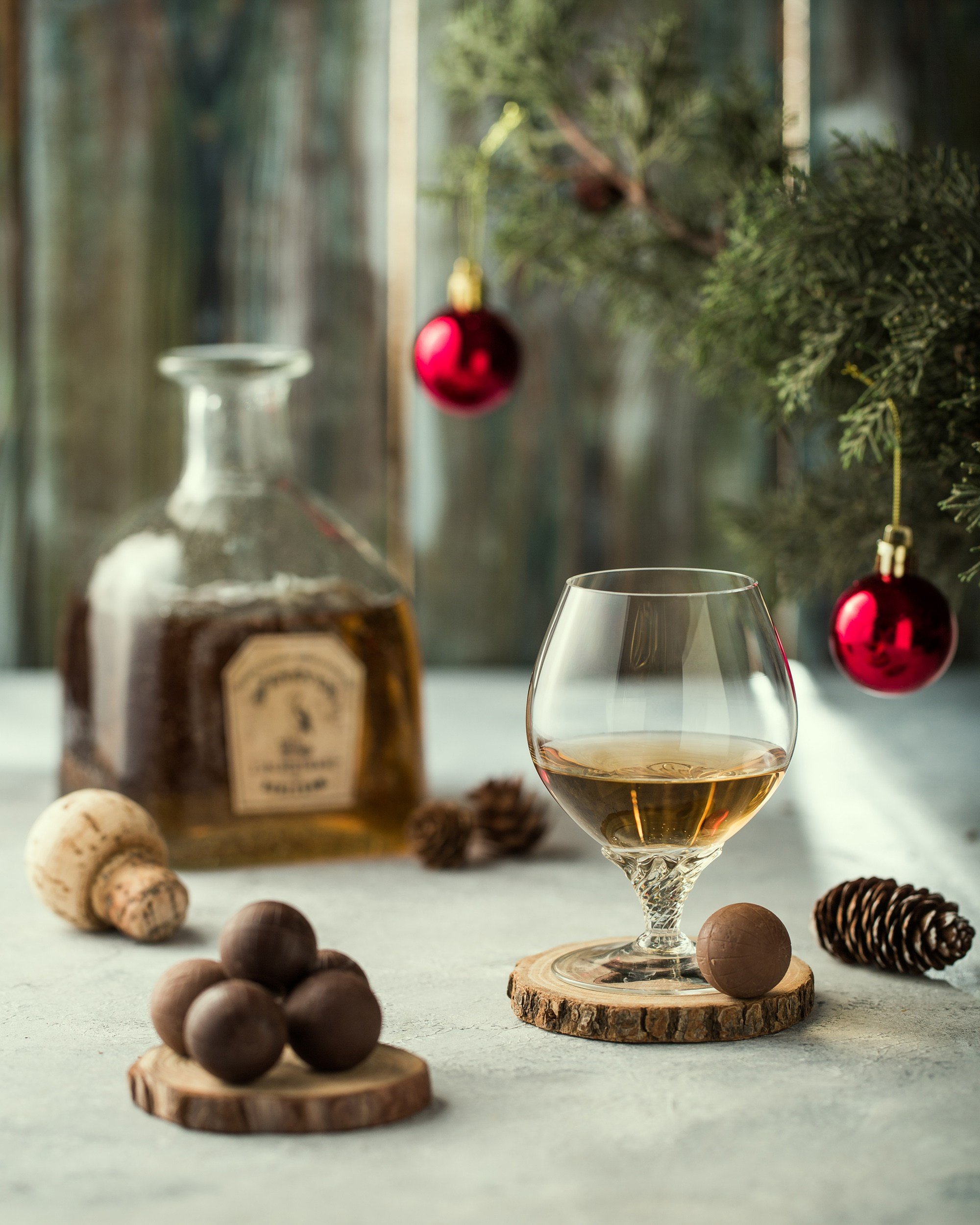 Boz daş səth üzərində, şam ağacı və qırmızı Milad şarları fonunda konyak/viski butulkası, mantar və şam qozası bəzəkləri. Ön planda taxta altlıq üzərində tək şokolad topu və konyak qədəhi, sol tərəfdə isə bir neçə şokolad topu.