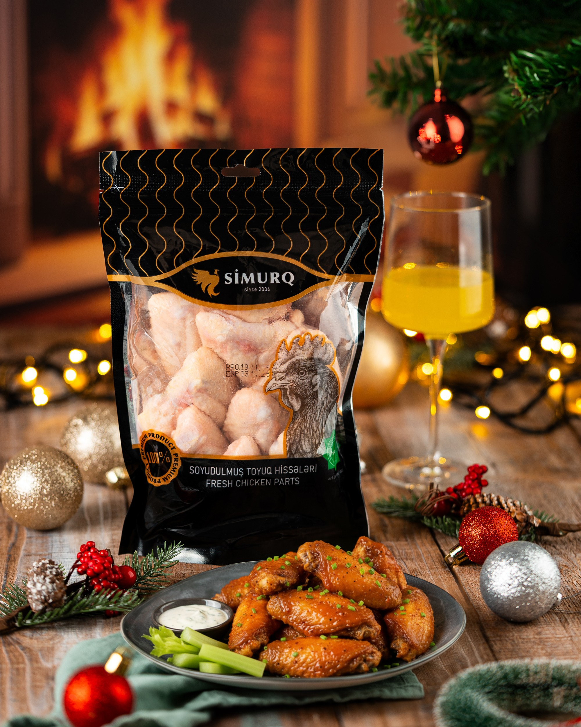 Şömine fonunda Milad işıqları ilə bəzədilmiş taxta səth üzərində, "SİMURQ" markalı soyudulmuş toyuq hissələri (qanadlar) paketinin önündə mavi boşqabda souslu qızardılmış toyuq qanadları. Sağda bir stəkan sarı içki görünür.