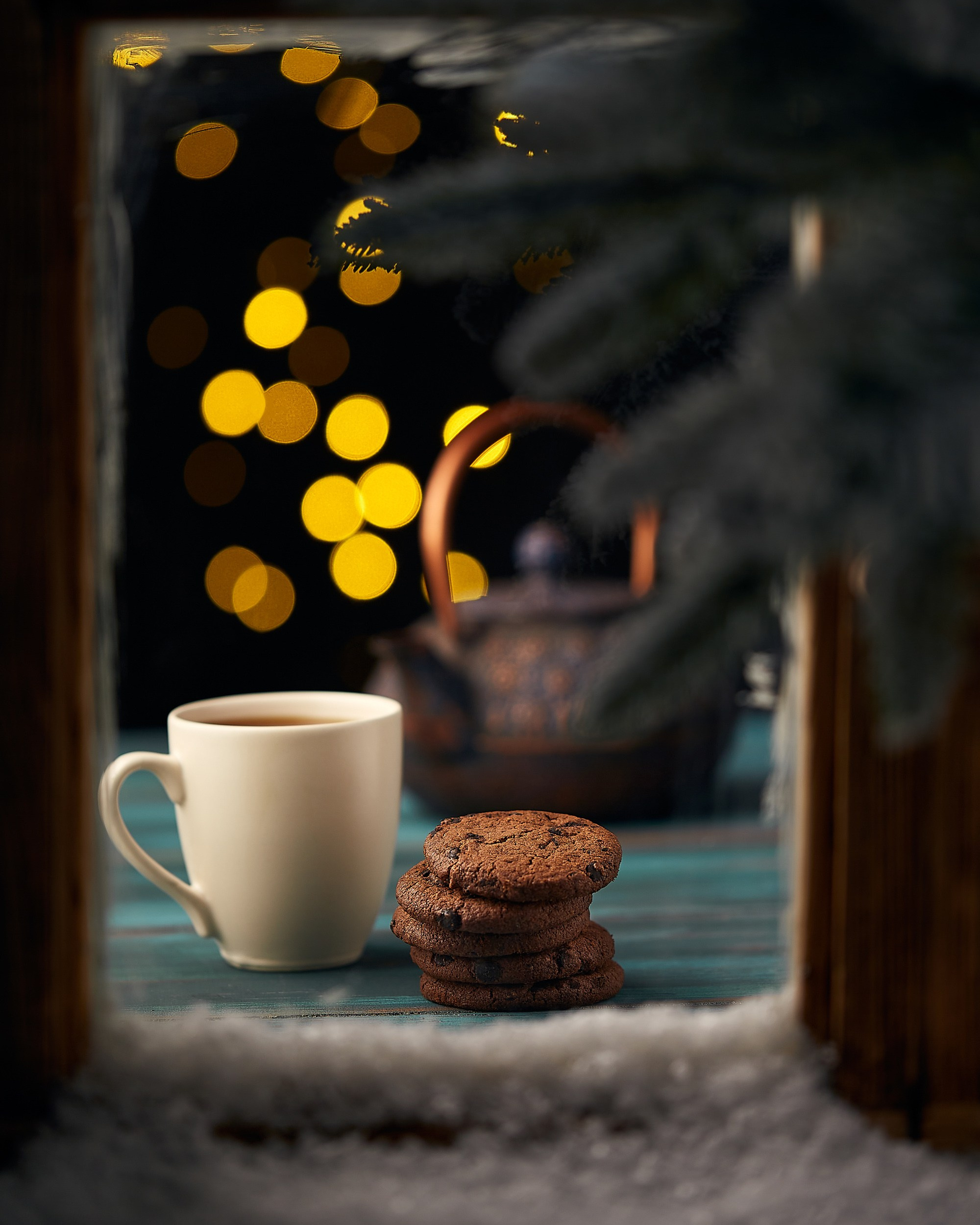 Süni qar ilə haşiyələnmiş pəncərədən qaranlıq fona baxış. Ön planda mavi taxta səth üzərində bir fincan çay və şokoladlı peçenyelər. Arxada bulanıq qızılı işıqlar və tünd rəngli çini çaydan görünür.