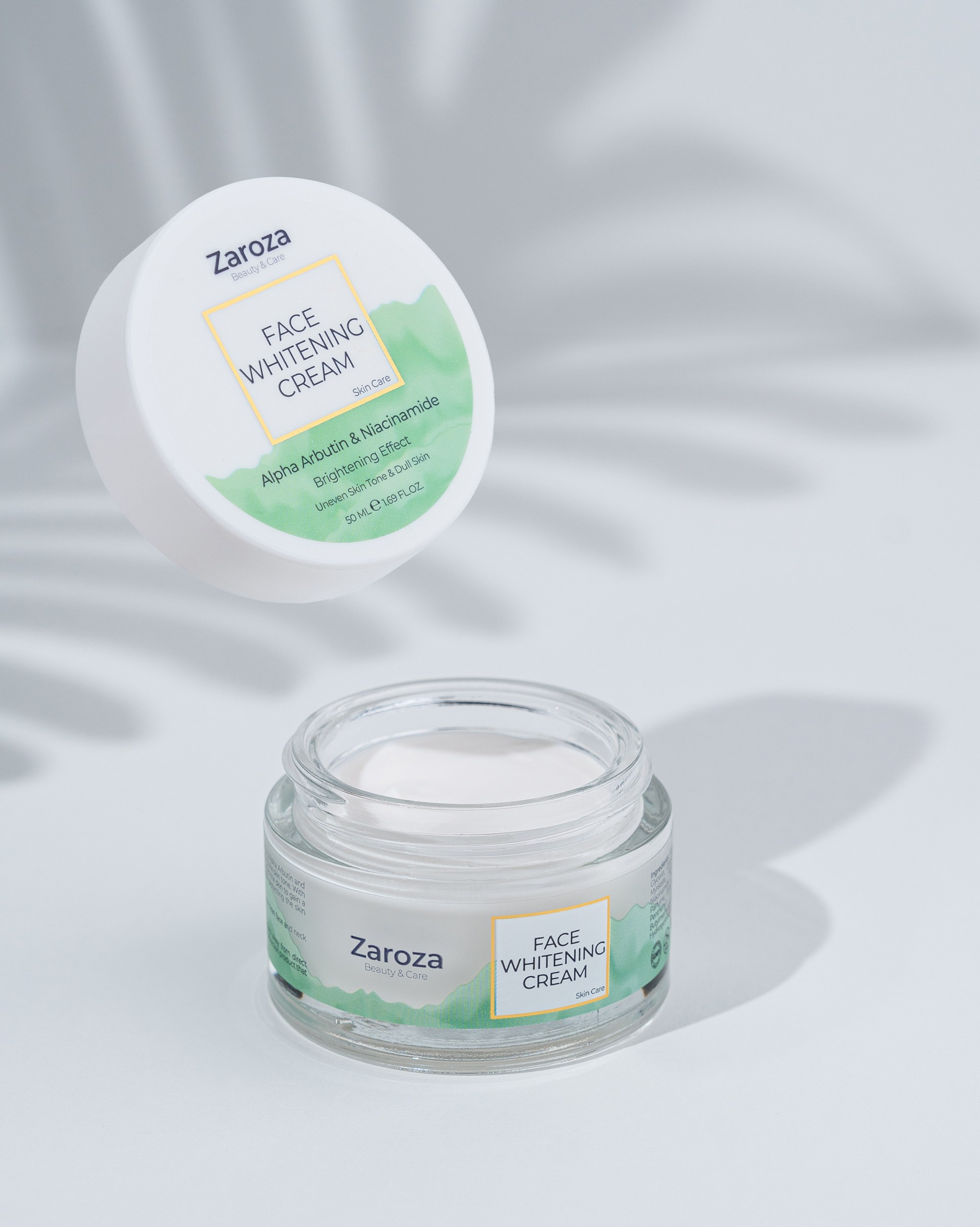 Açıq boz fonda havada uçan və masanın üzərində dayanan iki ədəd Zaroza Üz Ağardıcı Krem. Hər ikisi ağ qapaqlı şəffaf bankalardadır, üzərində "Face Whitening Cream" yazısı və yaşıl detallar görünür.