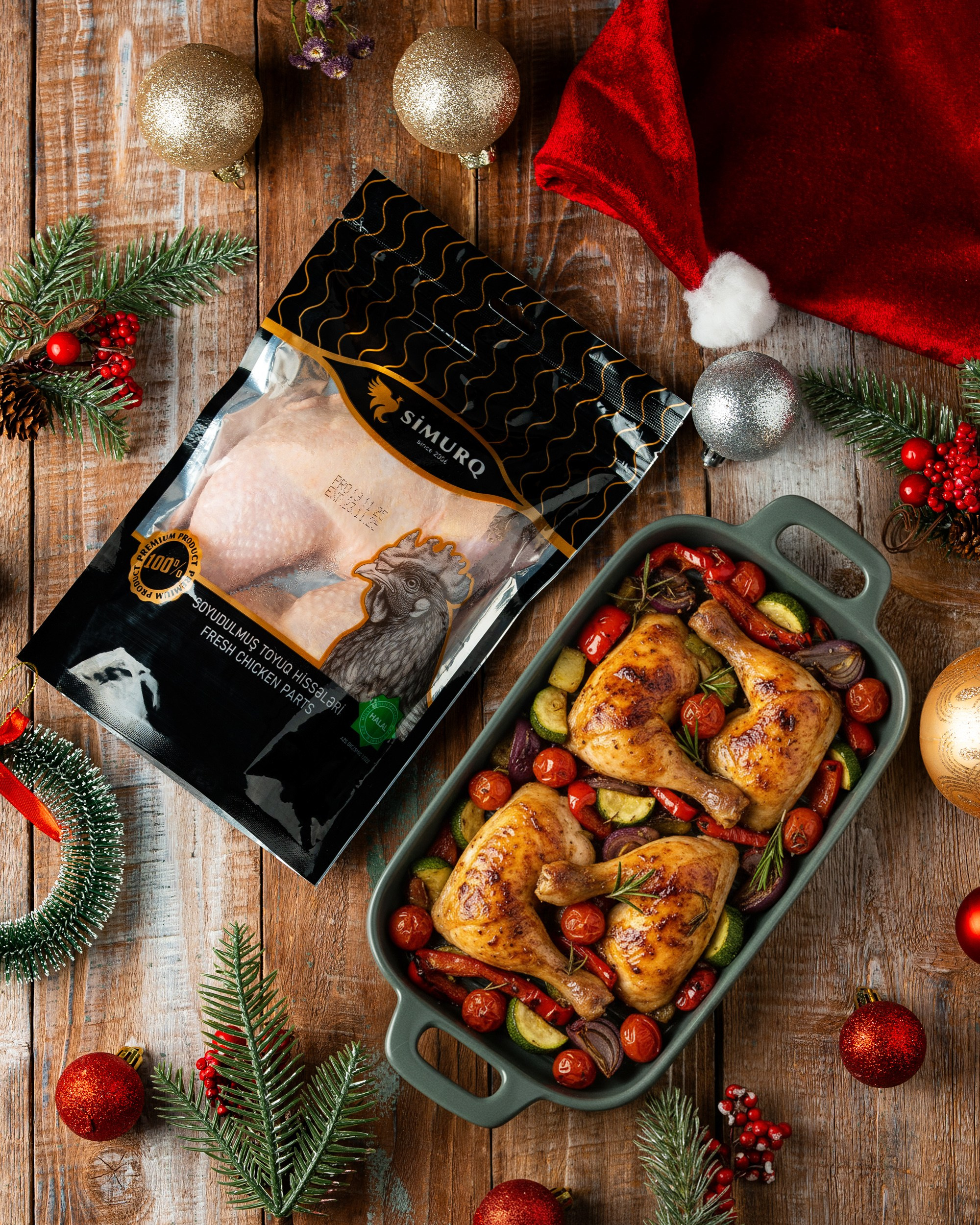 Taxta fon üzərində, şam bəzəkləri və Santa papağı yanında "SİMURQ" markalı soyudulmuş toyuq hissələri paketinin yanında tərəvəzlərlə (pomidor, bibər və s.) sobada bişirilmiş iki toyuq bud-bütün hissəsi olan boz qab.