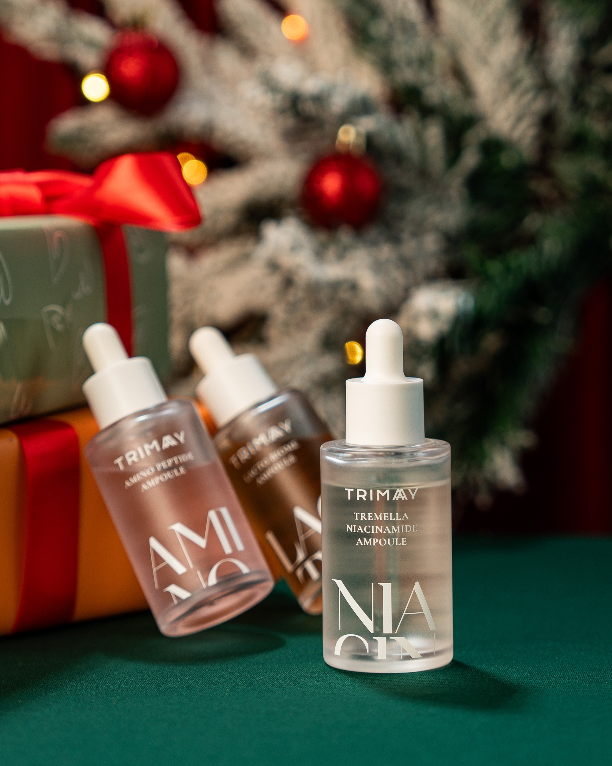 Yaşıl səth üzərində, Milad bəzəkləri və qırmızı lentli hədiyyələr fonunda Trimay markalı üç serum/ampula butulkası (Aminopeptid, Skvalan, Tremerla Niasinamid)