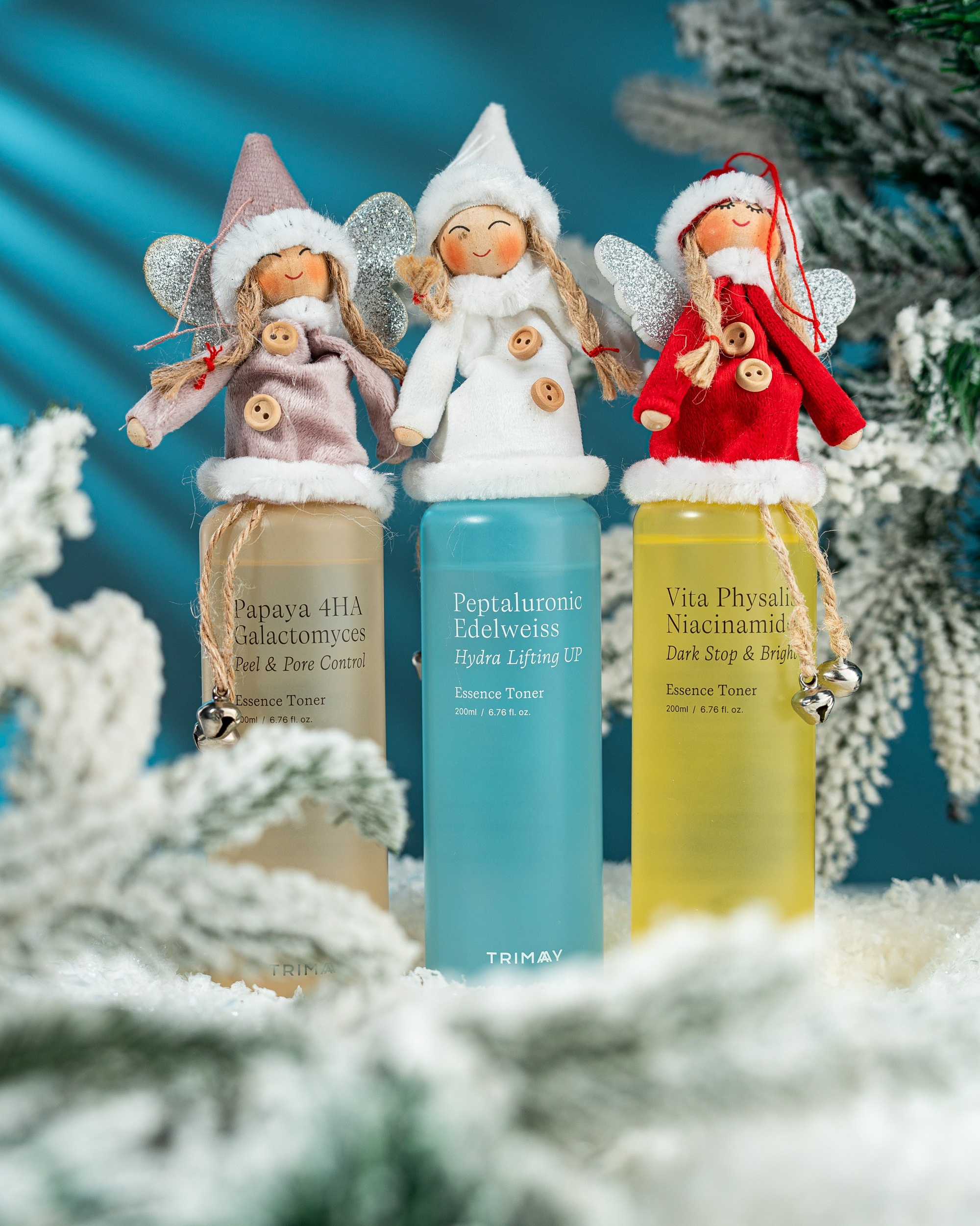 Qar və şam budaqları ilə bəzədilmiş qış mühitində Trimay Essence Toner butulkaları (Papaya 4HA, Peptaluronic Edelweiss, Vita Physalis Niacinamide). Hər butulkanın qapağında kiçik, Milad ruhlu mələk kuklaları dayanıb.