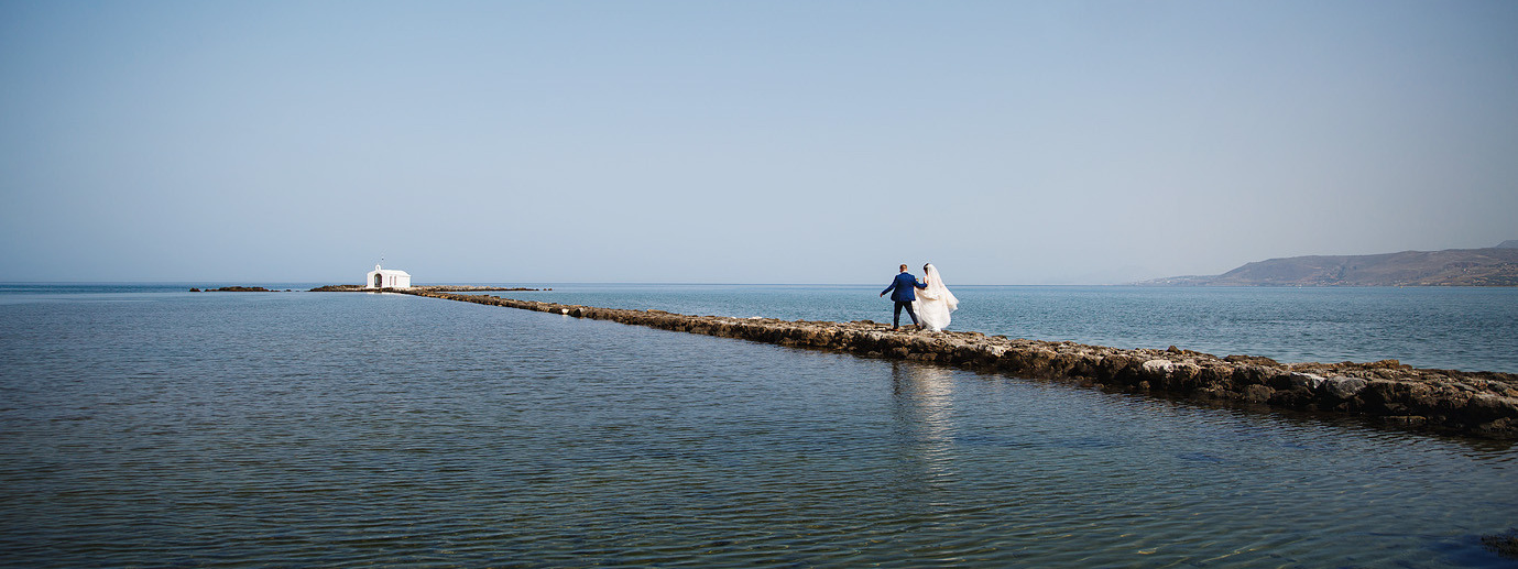 Молодожены идут по дорожке, выложенной из камней в море к часовне на Крите. Фотосессия в Греции.