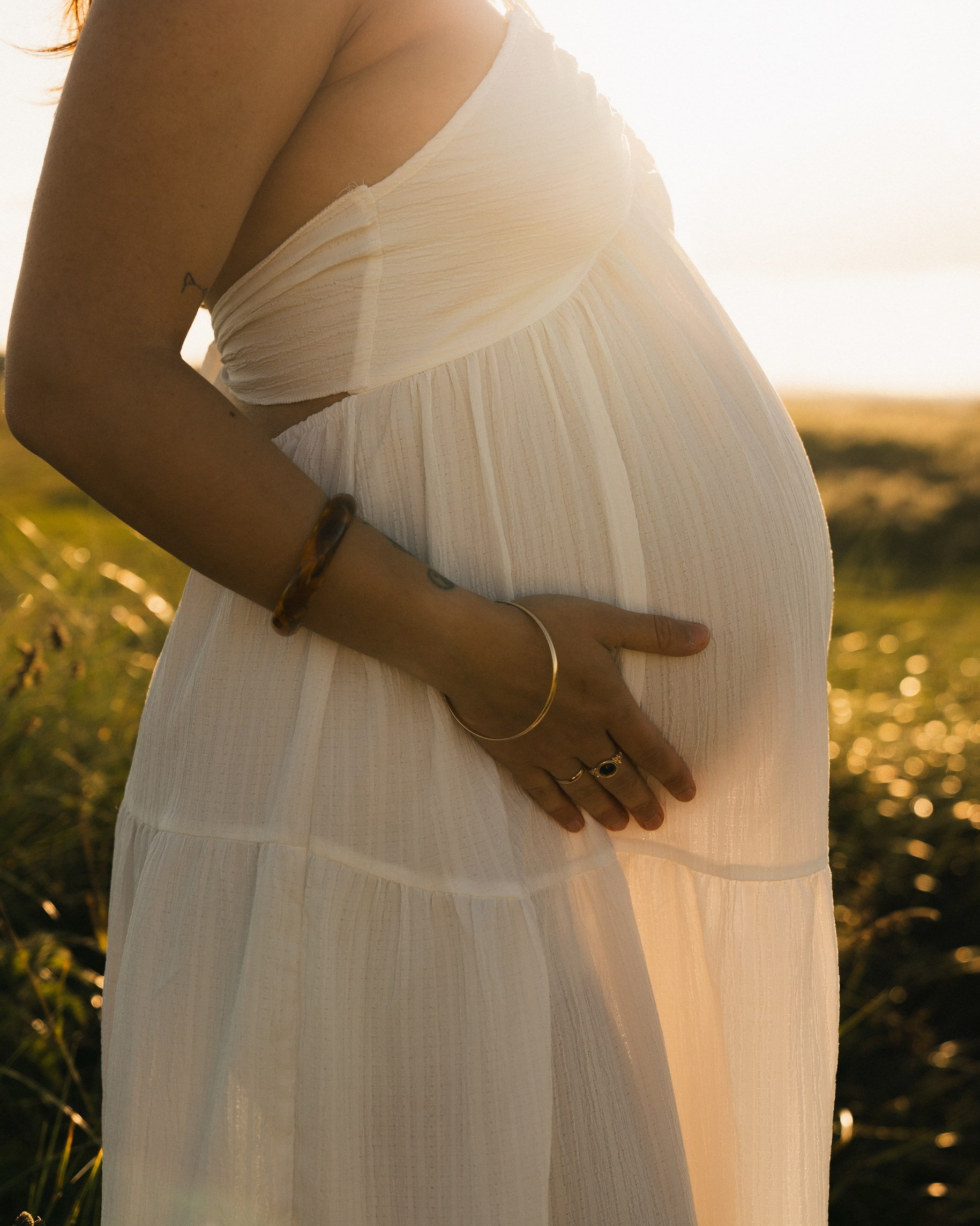 Close-up of pregnant woman in white dress holding her baby bump at golden hour, with partner’s hands gently embracing her. Backlit by warm sunset light at Druridge Bay beach, Northumberland.
