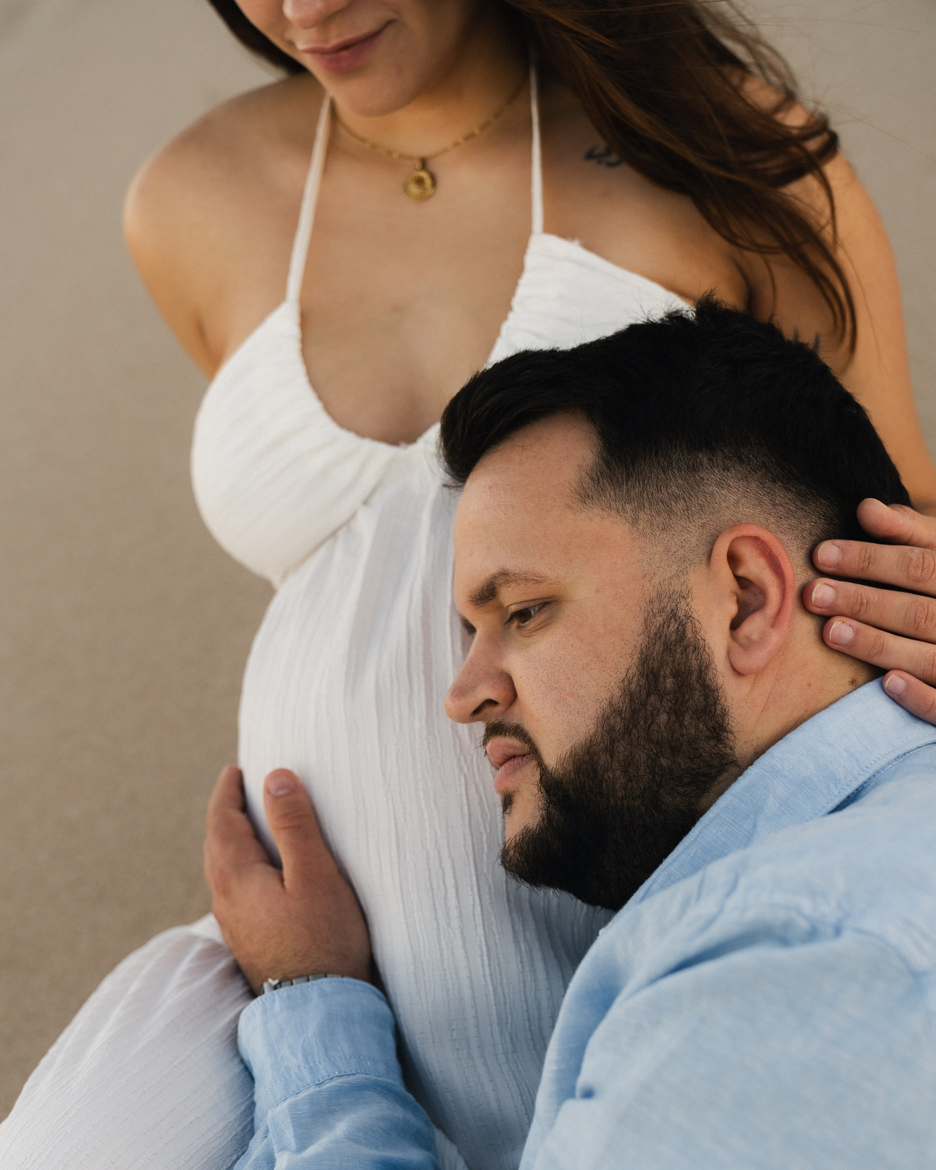 Expecting father embracing pregnant partner on the beach at Druridge Bay, Northumberland. Couple sharing intimate moment during golden hour maternity photo session in Newcastle.