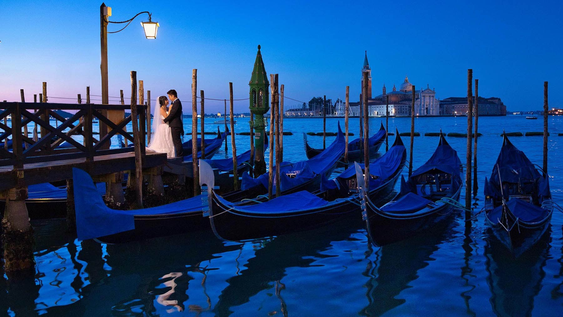 Newlyweds embrace under a gas lamp as gondolas float below them, during twilight in Venice - Kurt Vinion Photography