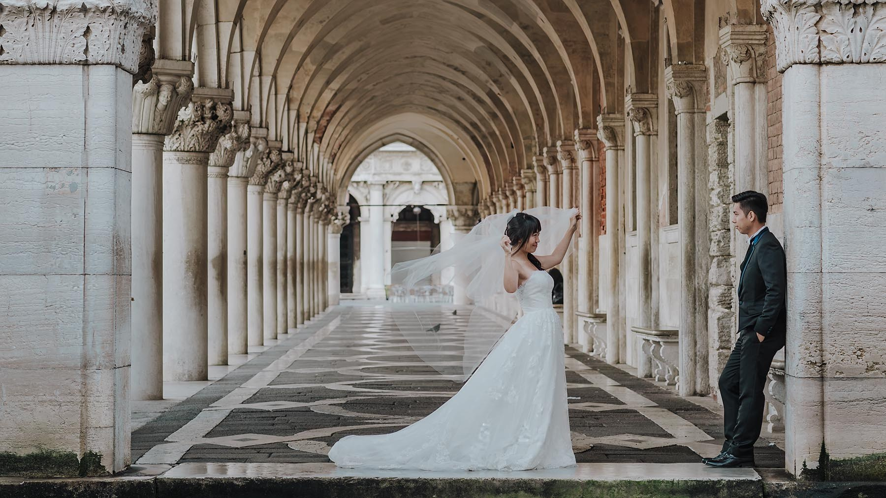 A bride plays with her veil as her groom watches under the historic arches of the Doge's Palace in Venice, Italy - Kurt Vinion Photography