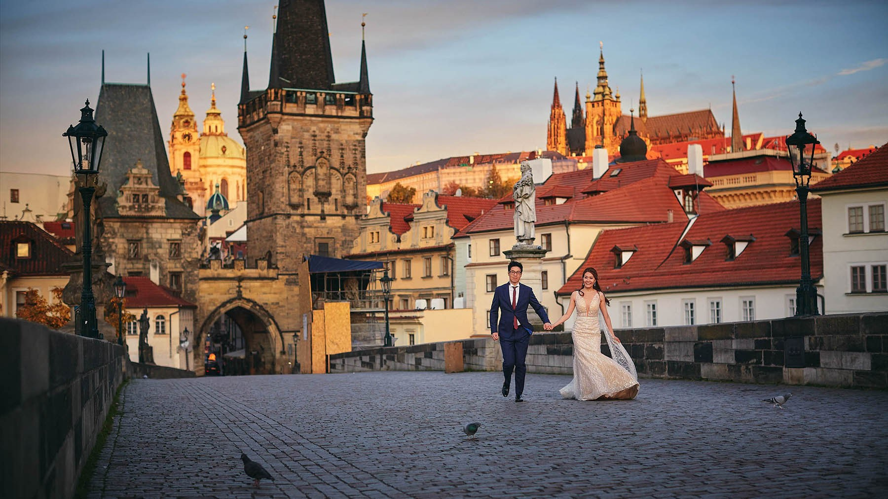 Couple walking on the empty Charles Bridge with illuminated Prague Castle at sunrise.
