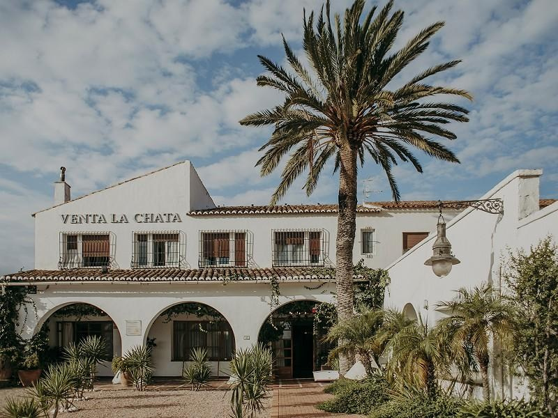 White façade and arched porch at Masía Venta La Chata wedding venue, with a tall palm tree in the courtyard.