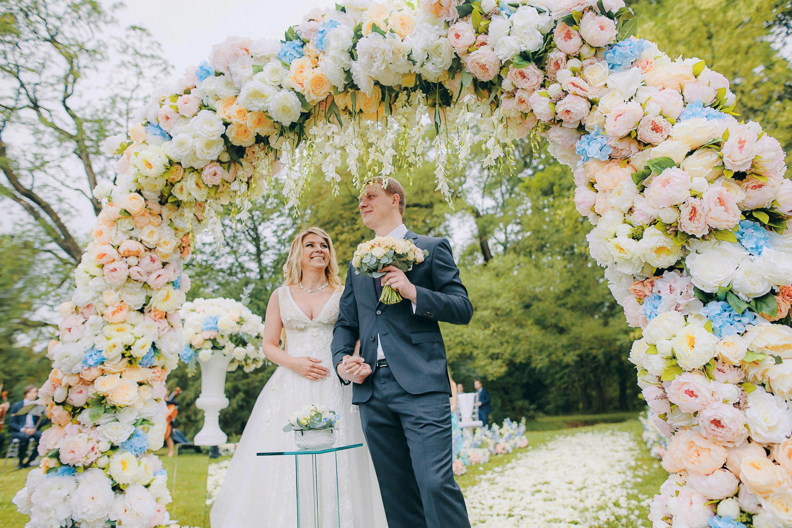 Beaming bride with proud groom holding bouquet under massive rose arch at castle wedding
