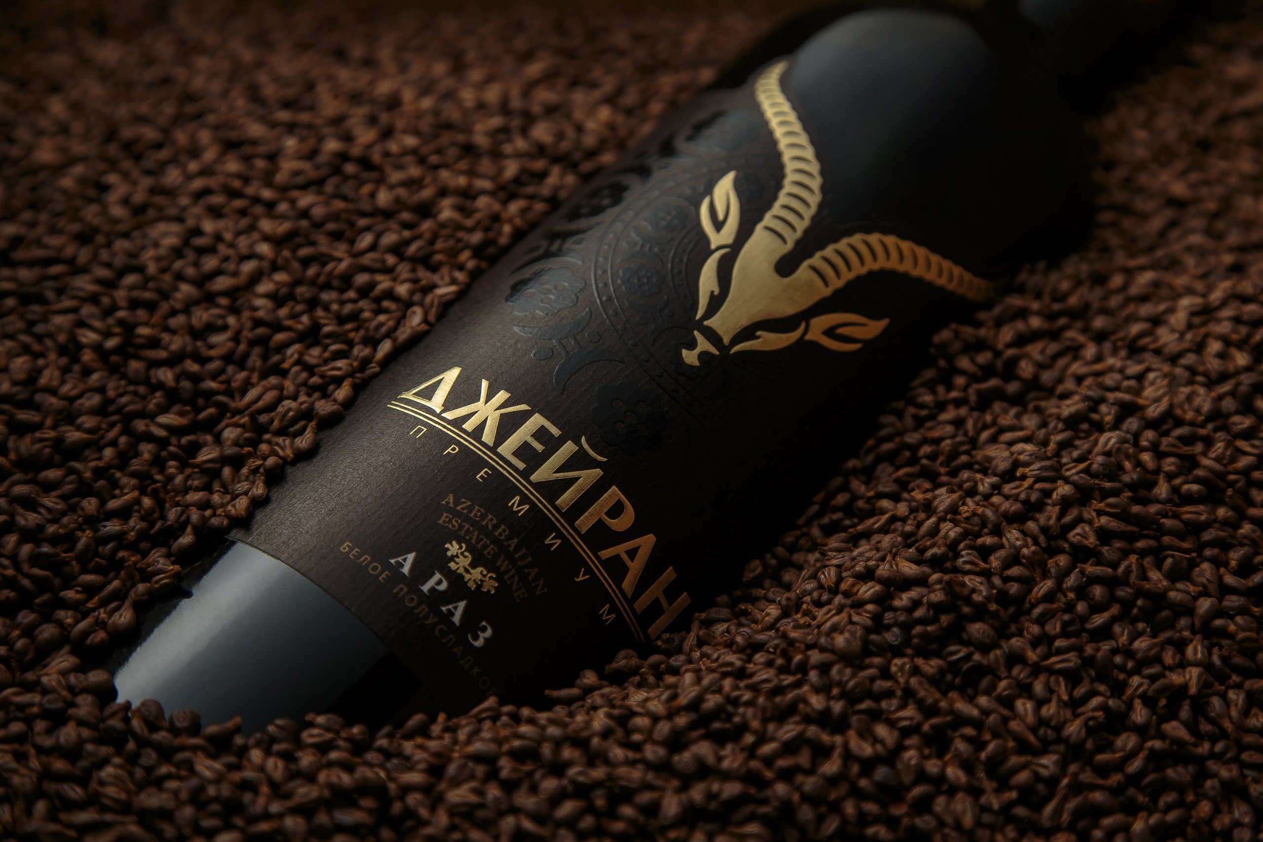 Ceyran Premium Azərbaycan şərabının qızılı ceyran loqosu olan qara şüşəsi, üzüm çəyirdəkləri (və ya qəhvə dənələri) içərisində yaxın plan.