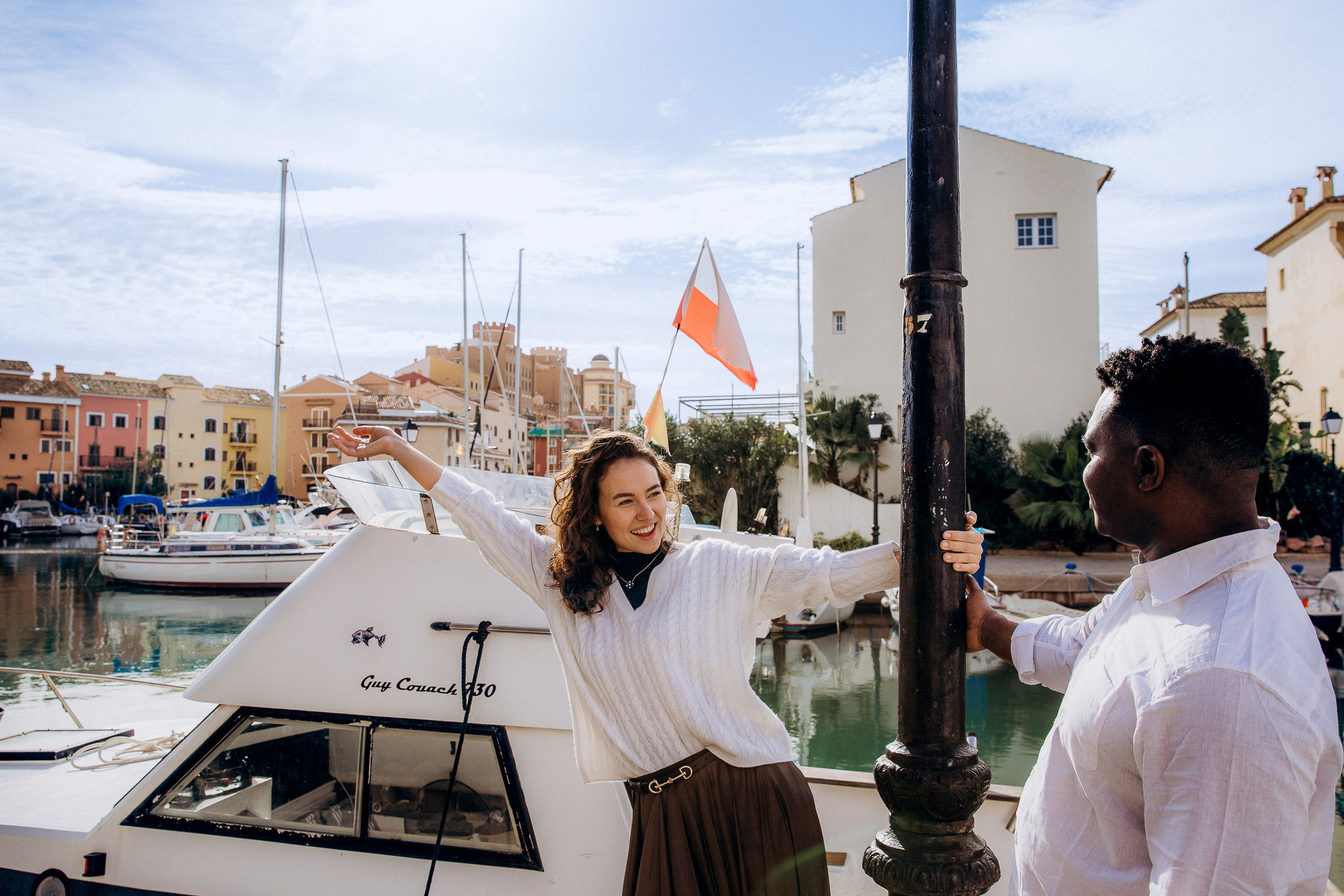Woman raising her hands to her head as her partner looks on beside the boats at Port Saplaya marina.