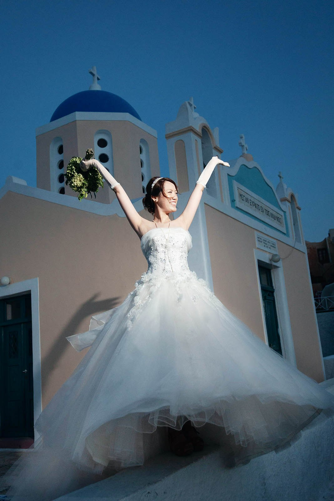 Bride in wedding dress raising her arms in joy with blue domes in the background, Oia Santorini.