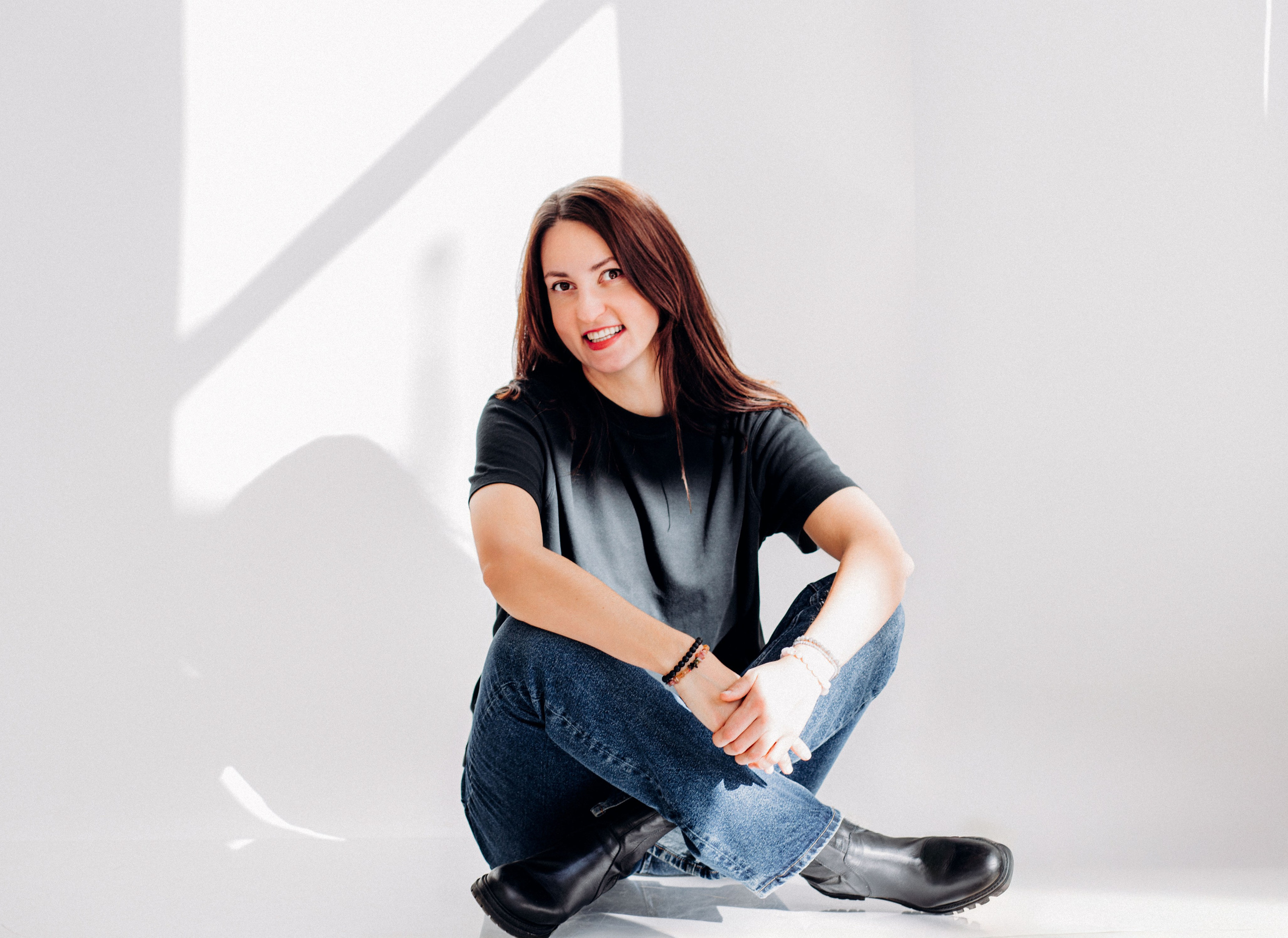 Retrato casual de una joven sonriente sentada con las piernas cruzadas en un estudio blanco minimalista con sombras de luz dramáticas, en Valencia, España.