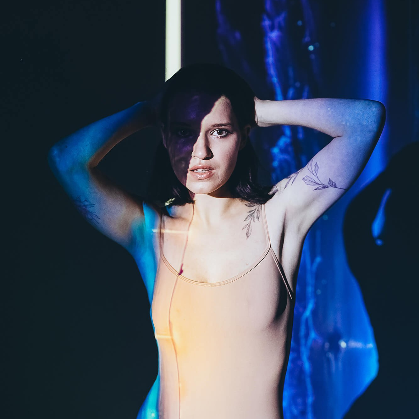 A bold and artistic portrait of a woman in a nude bodysuit, illuminated by dynamic blue and orange light projections. Her strong gaze and confident pose with hands behind her head symbolize self-acceptance, body positivity, and empowerment. The interplay of shadows and colors enhances the theme of transformation, inner strength, and embracing one's unique identity.