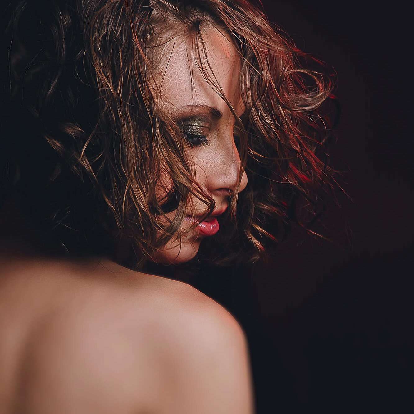 A sensual and intimate close-up portrait of a woman with tousled curly hair, her eyes closed and lips slightly parted. The soft lighting accentuates her bare shoulder and glowing skin, evoking themes of self-love, confidence, and empowerment. The dark background with a hint of red light adds a dramatic and artistic touch, enhancing the mood of self-expression and feminine strength.
