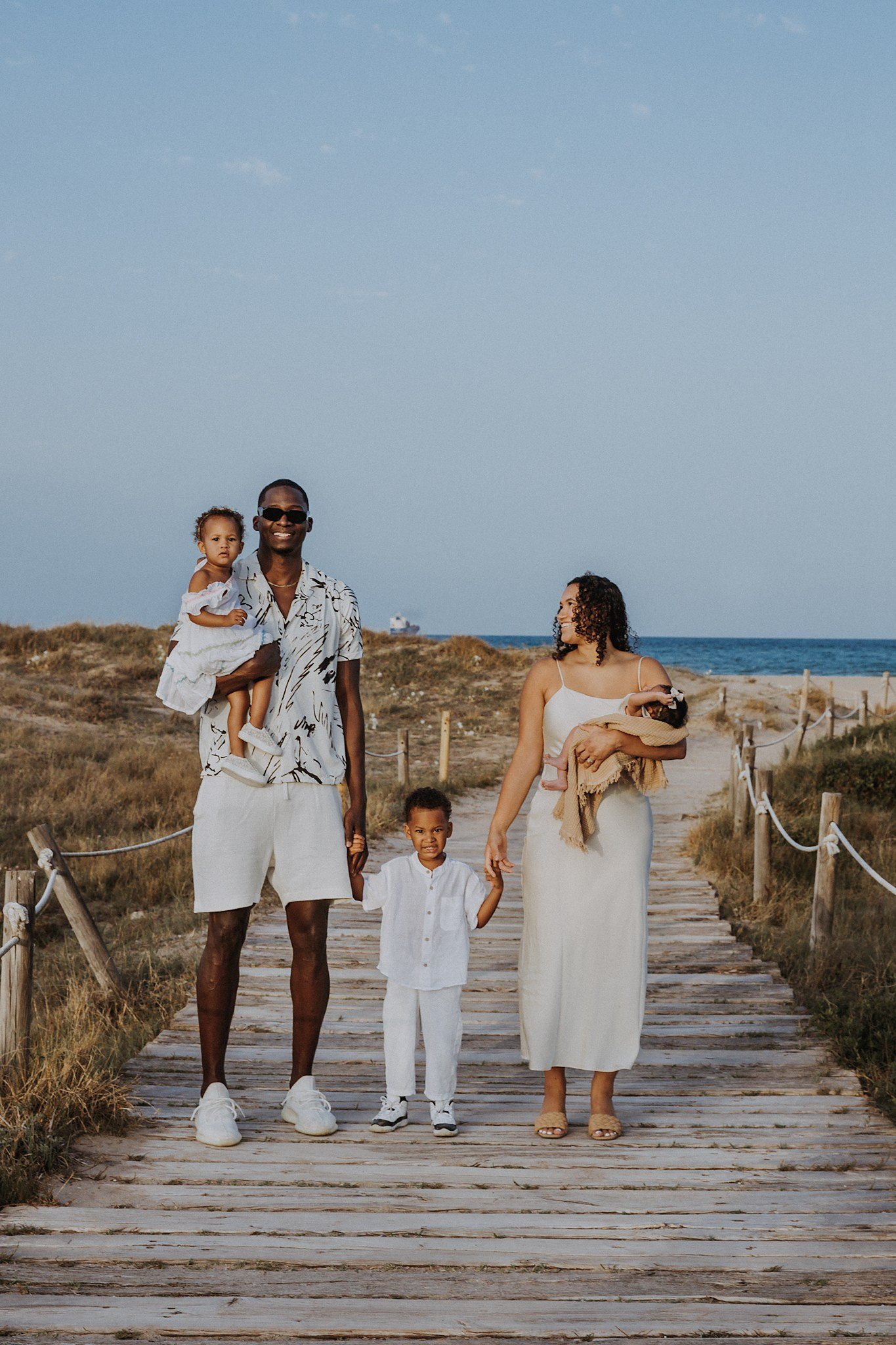 Hermosa familia caminando de la mano por un sendero de madera junto a la playa en Valencia, España — un momento entrañable capturado durante una sesión familiar al atardecer.