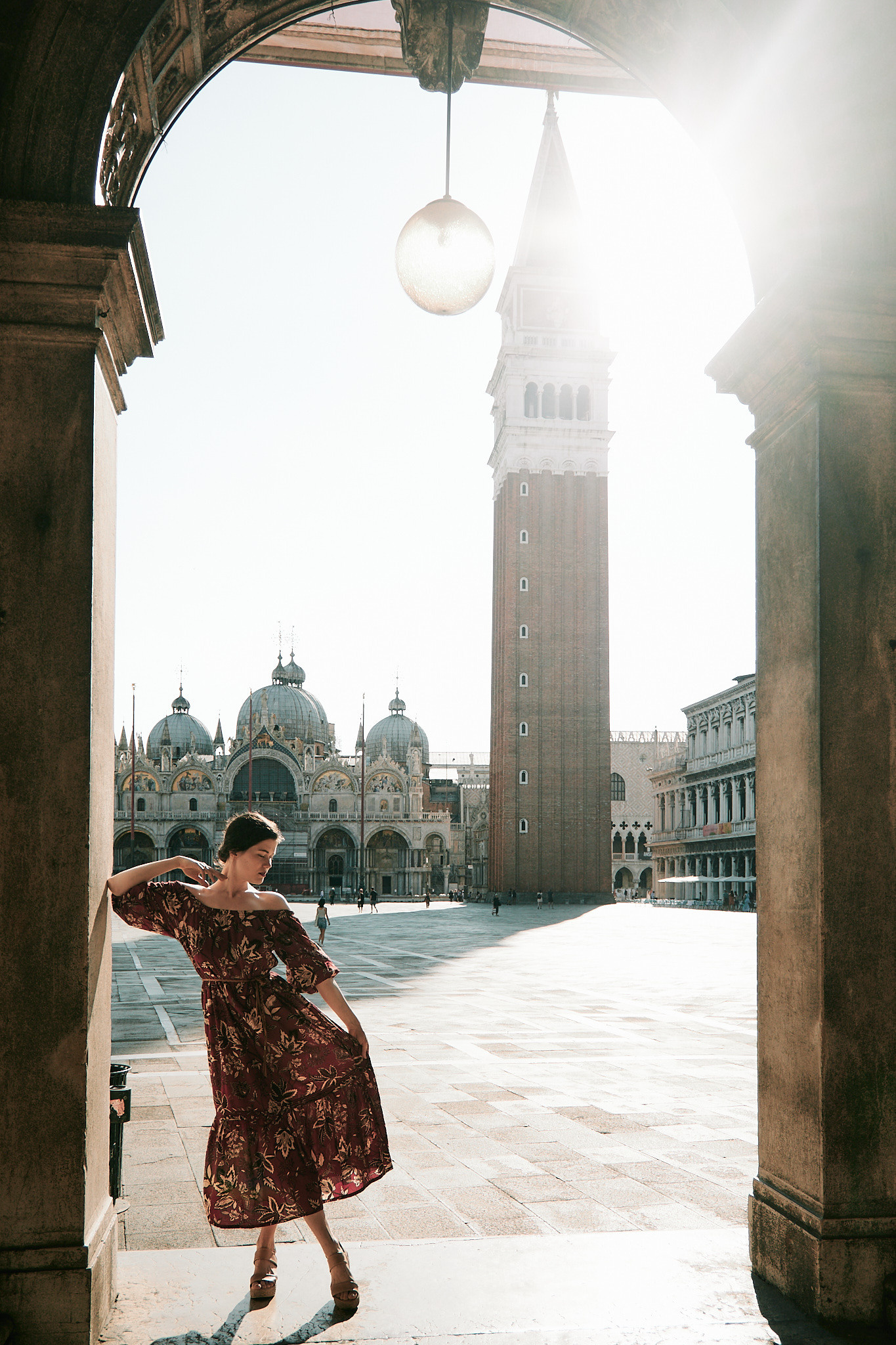A young woman stands on San Marco Square, gazing at the stunning architecture and bustling crowds around her. The sun casts a warm glow on her face, highlighting her delicate features and soft, flowing hair. She wears a flowing dress that seems to dance in the breeze, accentuating her graceful movements. The intricate details of the nearby buildings and the vibrant colors of the street performers create a romantic and captivating atmosphere around her, making for a beautiful and memorable portrait.