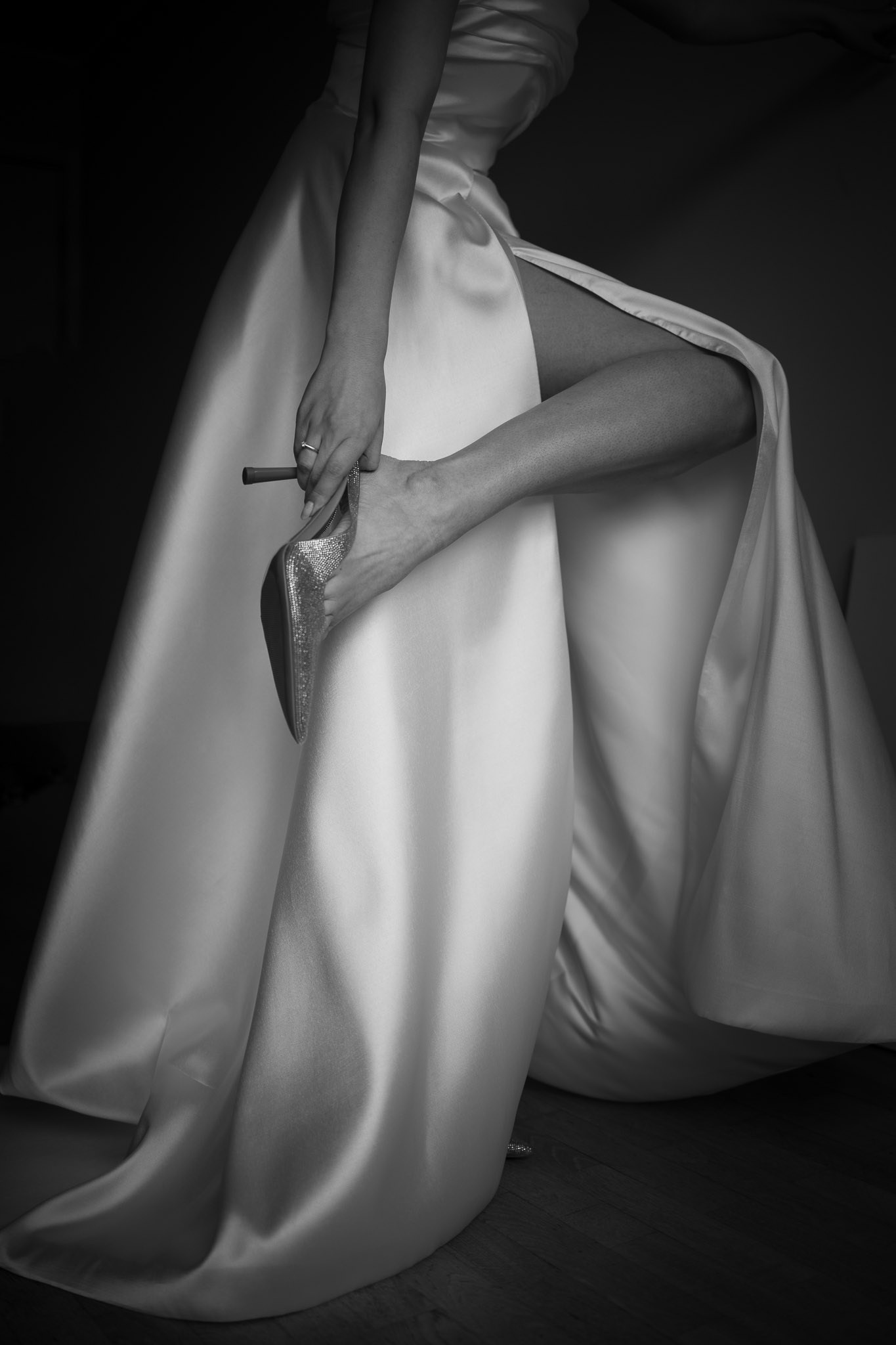 Crno-bela fotografija žene u elegantnoj satenskoj venčanici kako obuva svetlucave cipele sa visokom potpeticom. Njena ruka podešava cipelu, a na prstu se vidi prsten.