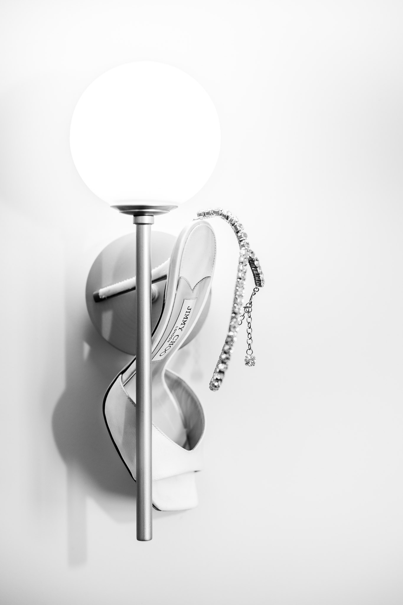 Minimalistička crno-bela slika koja prikazuje belu Jimmy Choo venčanu cipelu sa svetlucavim kristalima  pored zidne lampe.