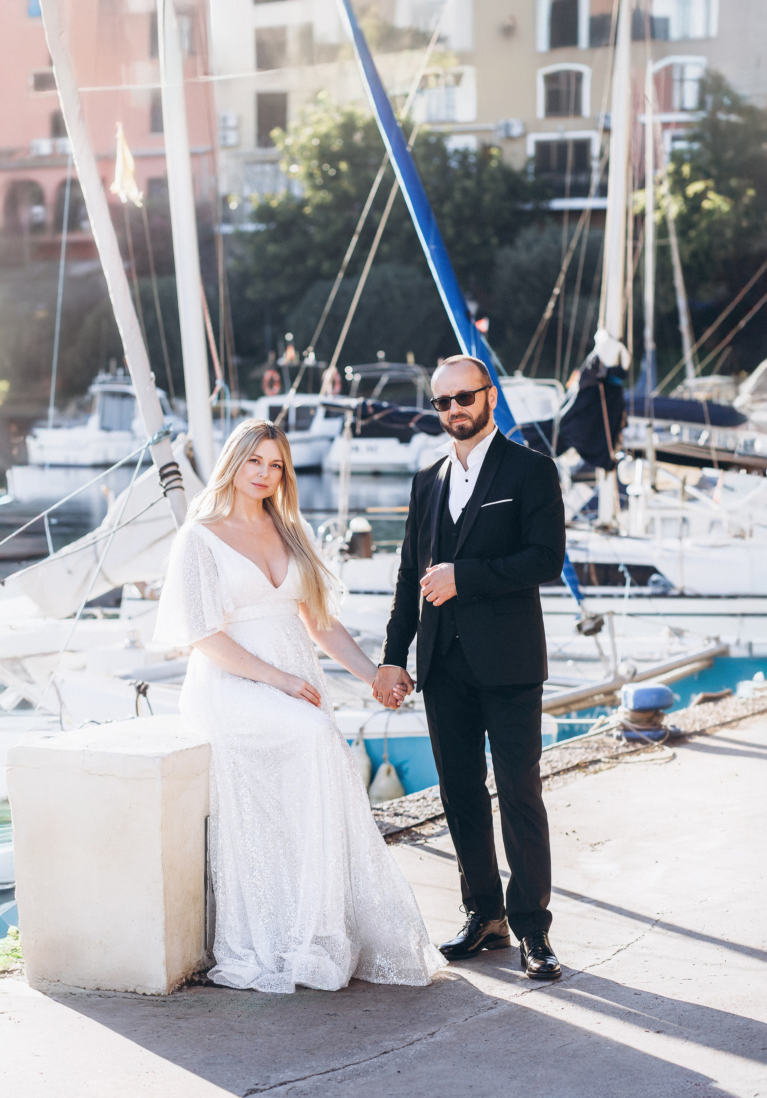 Woman in a long white dress holding hands with a man in a dark suit beside the boats at Port Saplaya marina.