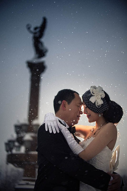 A couple embraces in the snow near a statue at the Rudolfinum in Prague during their winter pre-wedding.