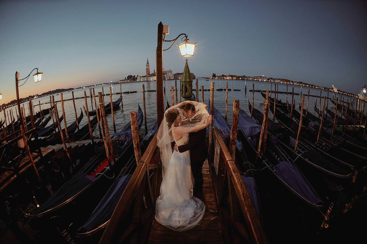 Bride and groom embrace under a gas lamp above the gondolas in Venice at night.