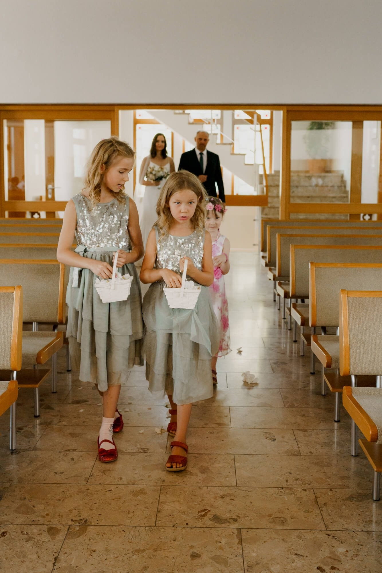 Two flower girls in matching sage green dresses walking down the aisle carrying baskets, guests seated on either side