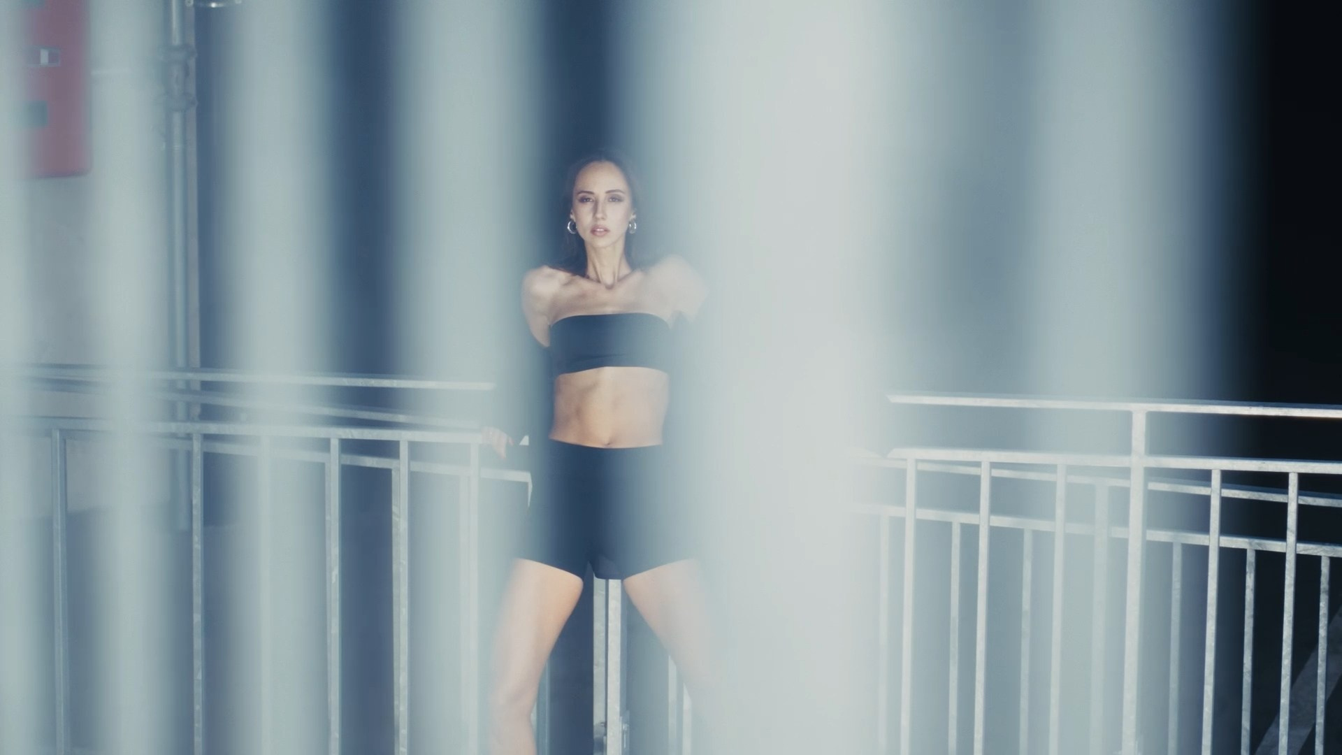 A distant view of a person leaning against a railing in a dimly lit indoor space, wearing athletic wear and exuding a calm and contemplative mood.