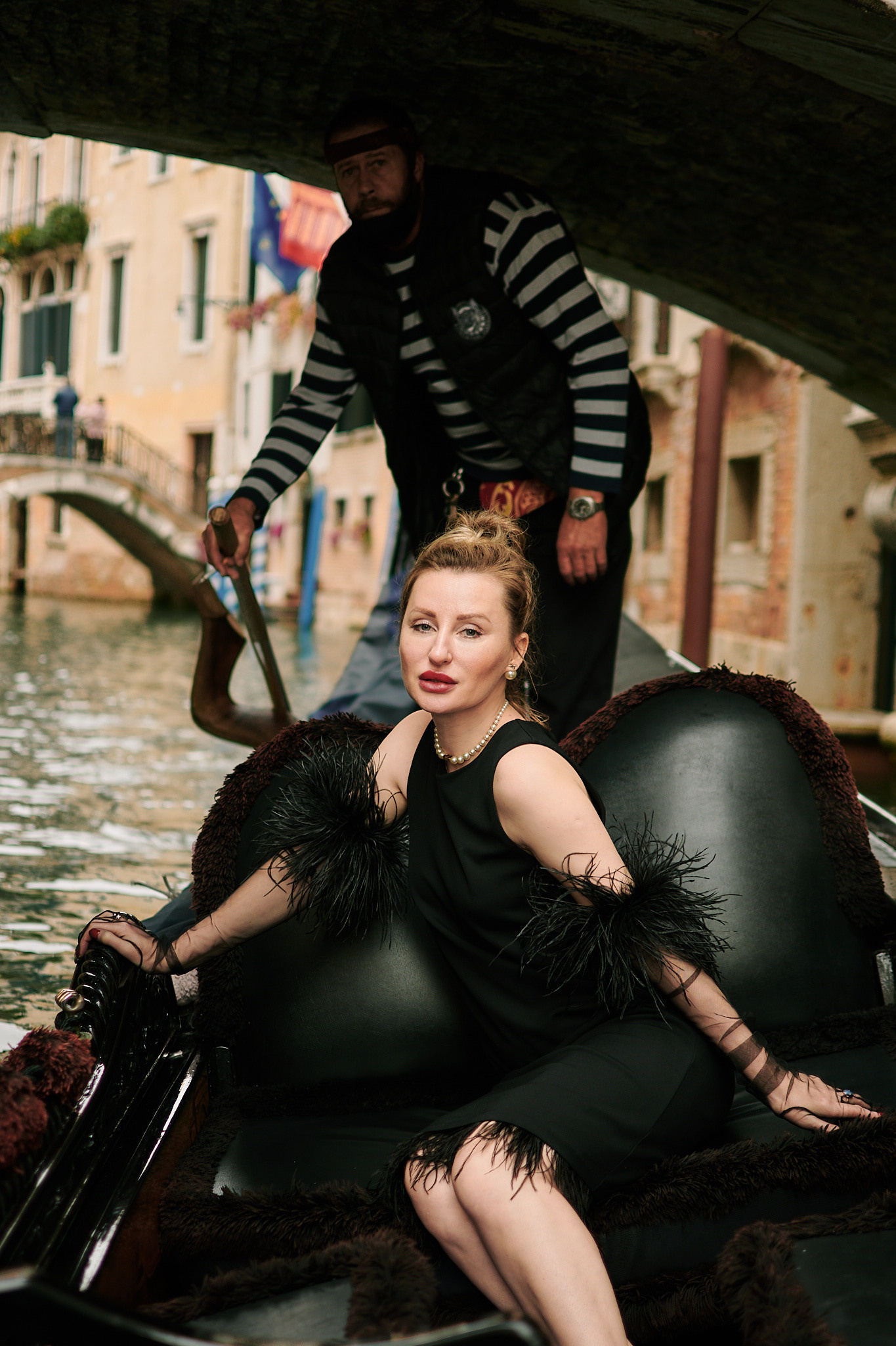 A mesmerizing young woman in a black dress sits on a gondola, gliding through the narrow streets of Venice. The black and white striped shirt of the gondolier is visible in the background. The woman's silky hair flows in the breeze, and her captivating eyes gaze off into the distance. The winding streets of Venice create a beautiful, romantic backdrop for this enchanting photo. Fashion blogger shoot Venice. Portrait and Fashion Photographer in Venice, Italy. Instagram Photos with lifestyle photographer
